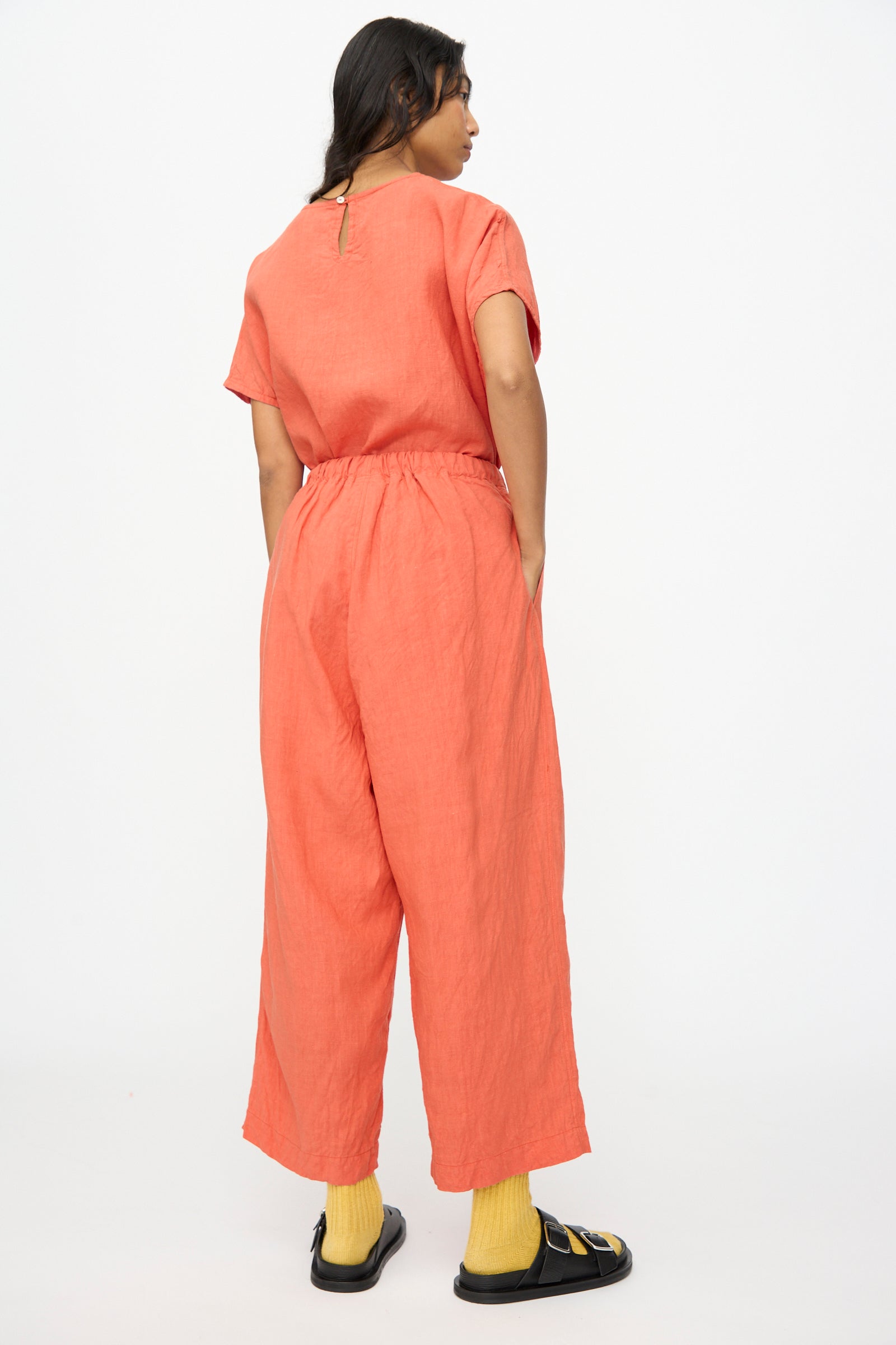 A person stands with their back to the camera wearing Ichi Antiquités' French Linen Pants in Orange, paired with yellow socks and black sandals, against a plain white background.