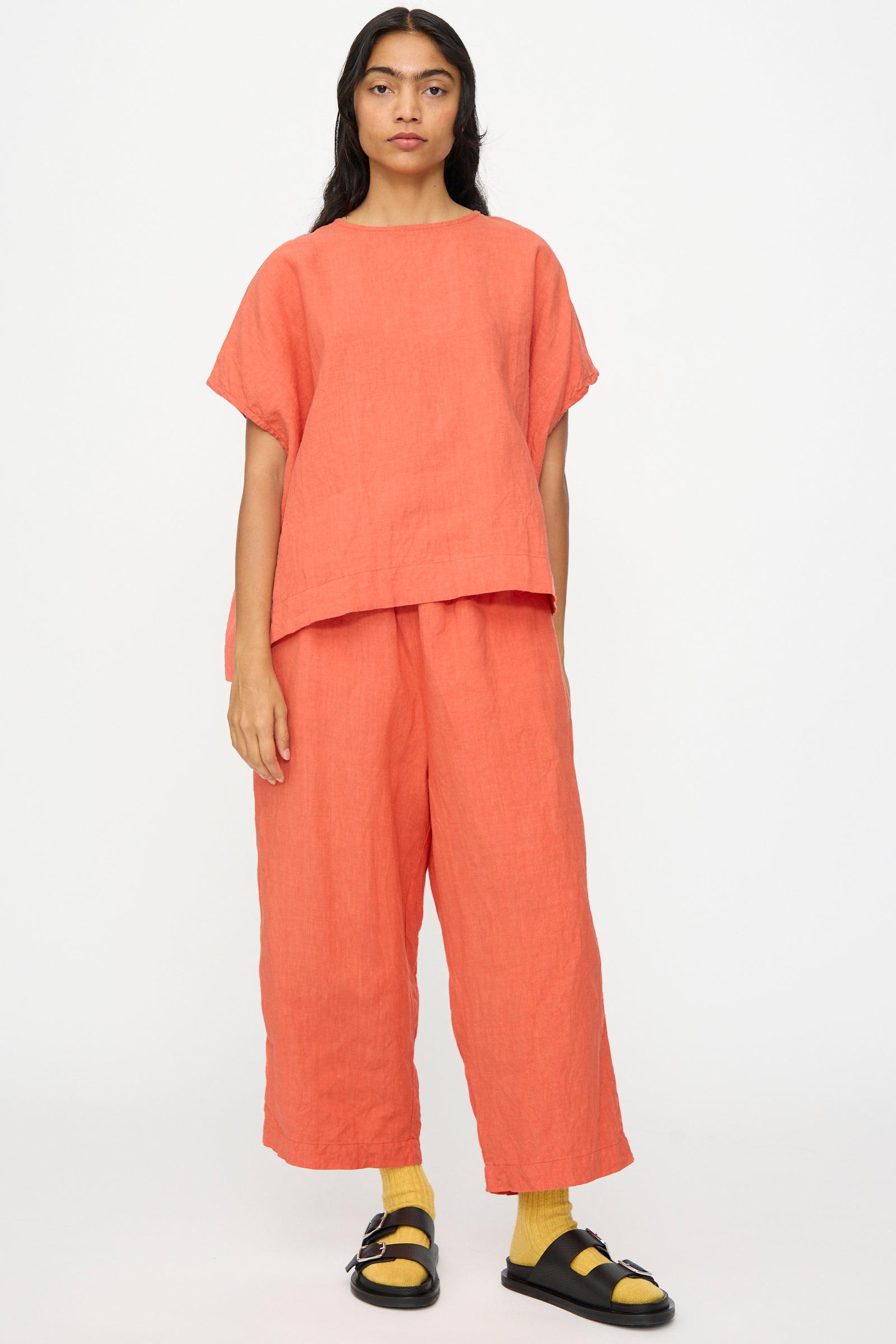 A person wears the Ichi Antiquités French Linen Pullover in Orange with matching wide-leg pants, yellow socks, and black sandals, standing against a plain white background.