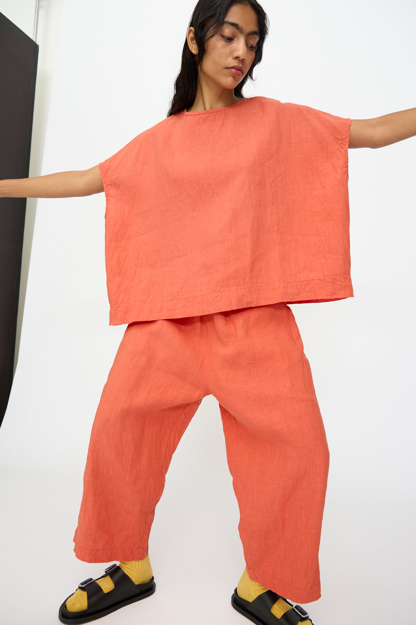 A person stands with arms outstretched in the Ichi Antiquités French Linen Pullover in Orange, paired with matching pants, yellow socks, and black sandals against a white background.