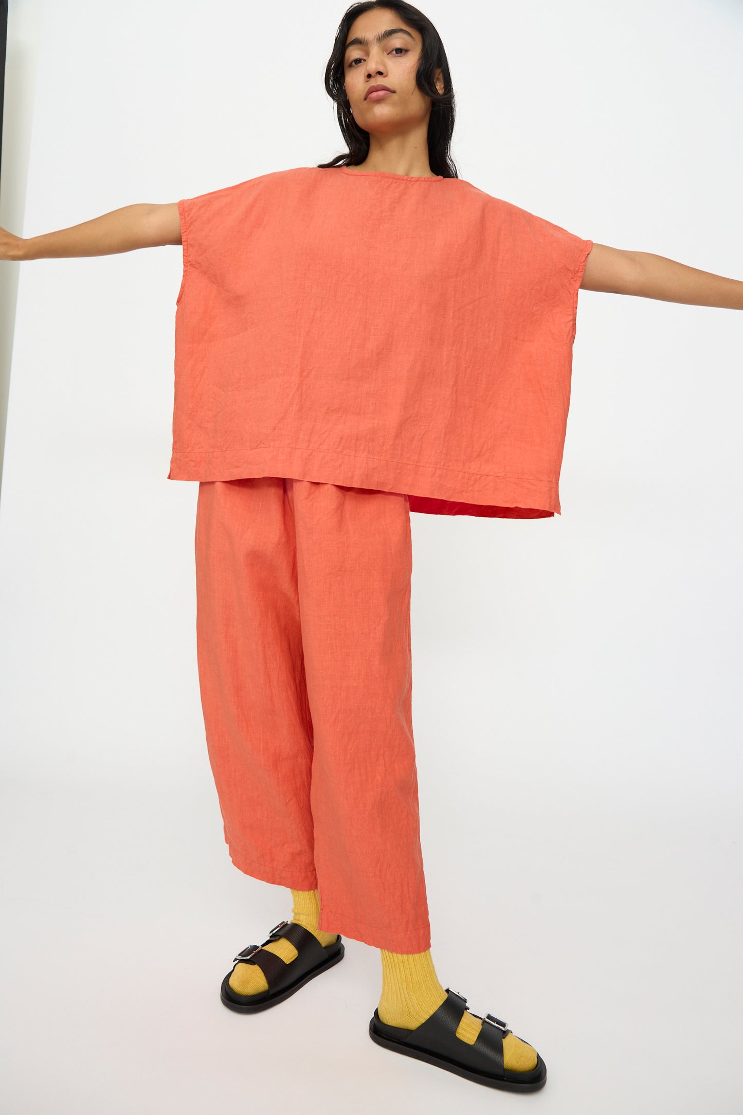 Person wearing a boxy fit Ichi Antiquités French Linen Pullover in Orange, matching pants, yellow socks, and black sandals stands with arms outstretched against a plain background.
