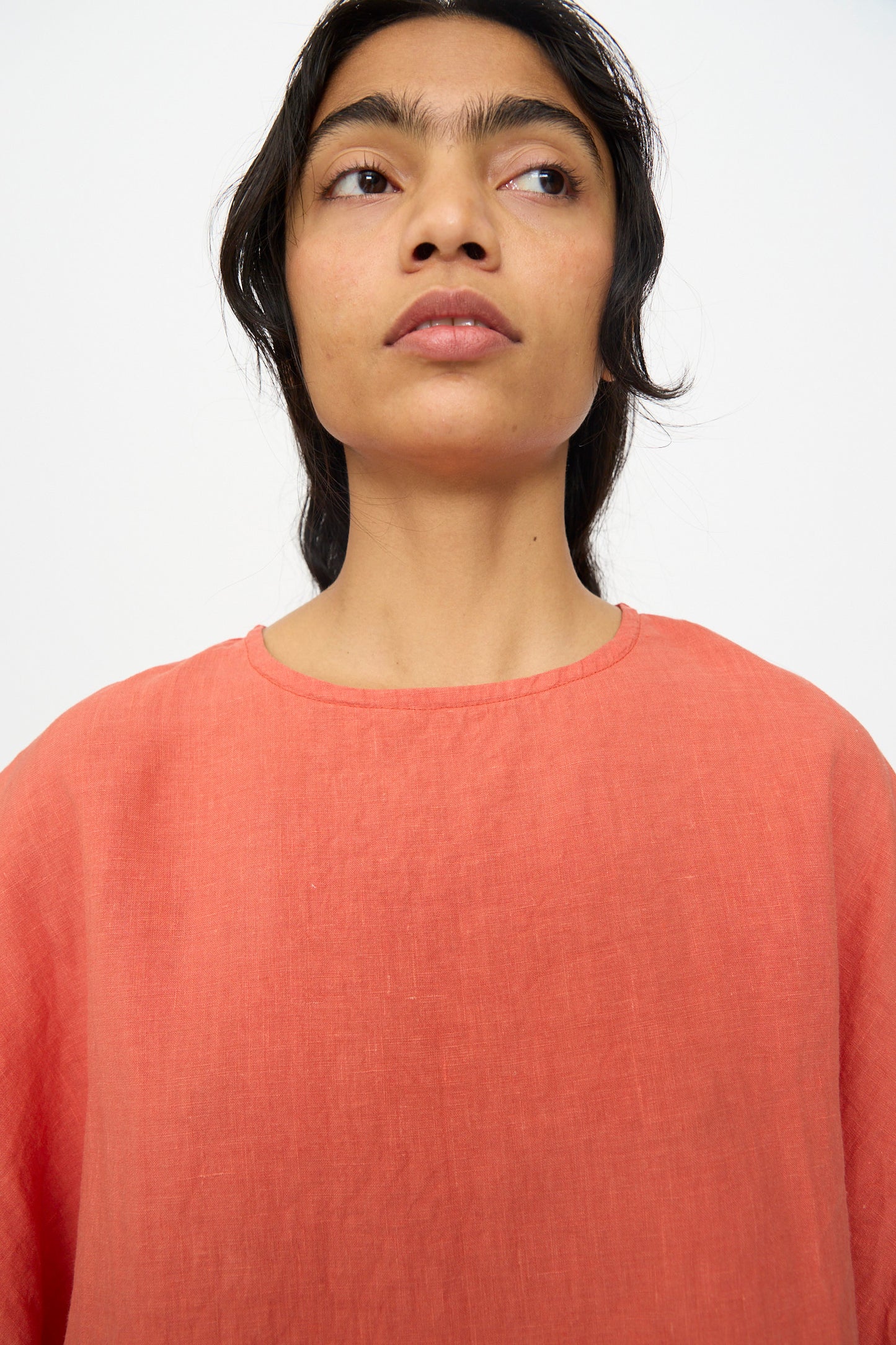 A person with long dark hair wears the Ichi Antiquités French Linen Pullover in Orange, featuring a boxy fit, while looking slightly upwards against a plain white background.