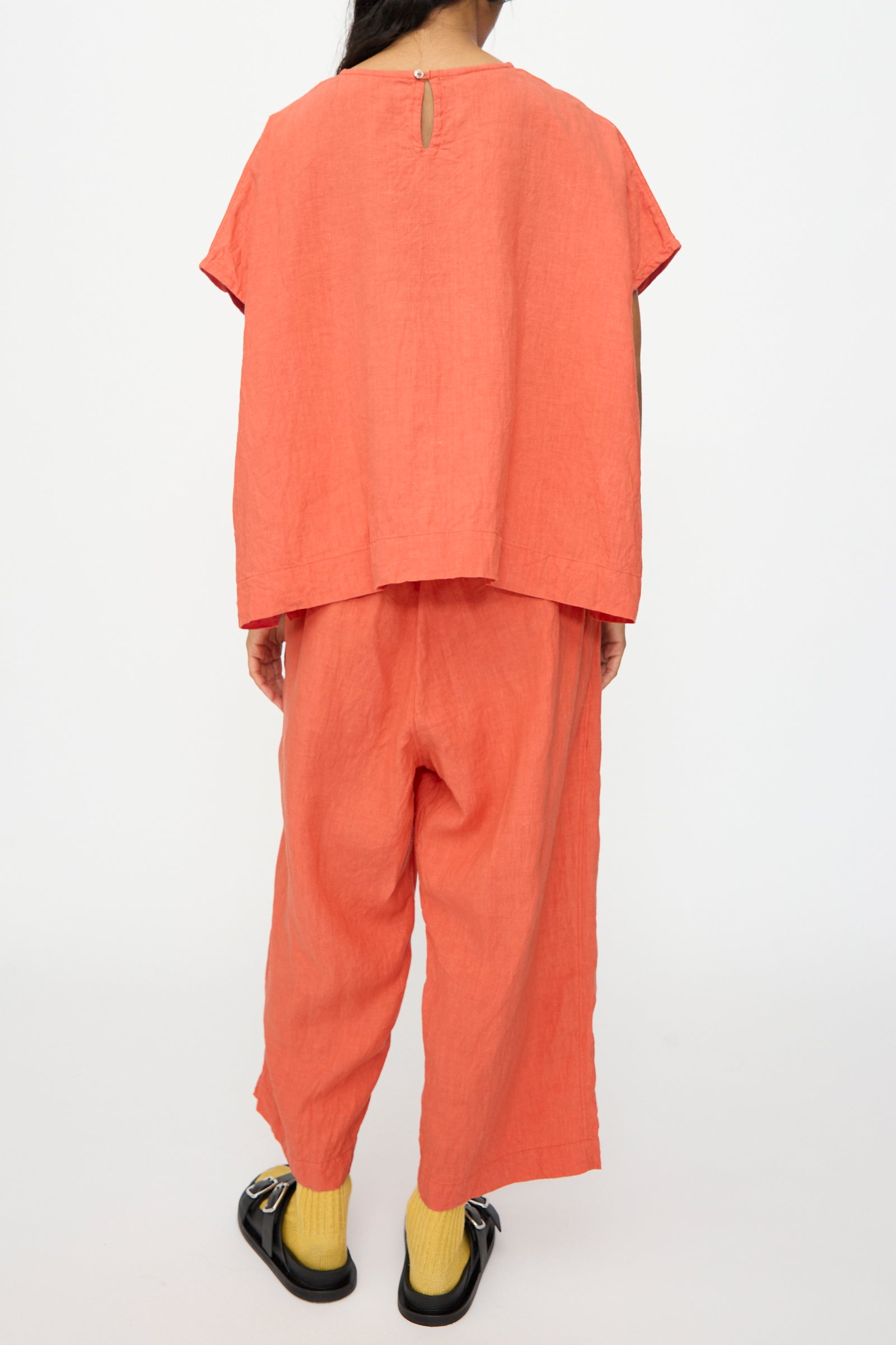 A person stands with their back to the camera in a boxy fit French Linen Pullover in Orange by Ichi Antiquités, paired with pants, yellow socks, and black sandals against a plain background. This relaxed outfit is made in Japan.