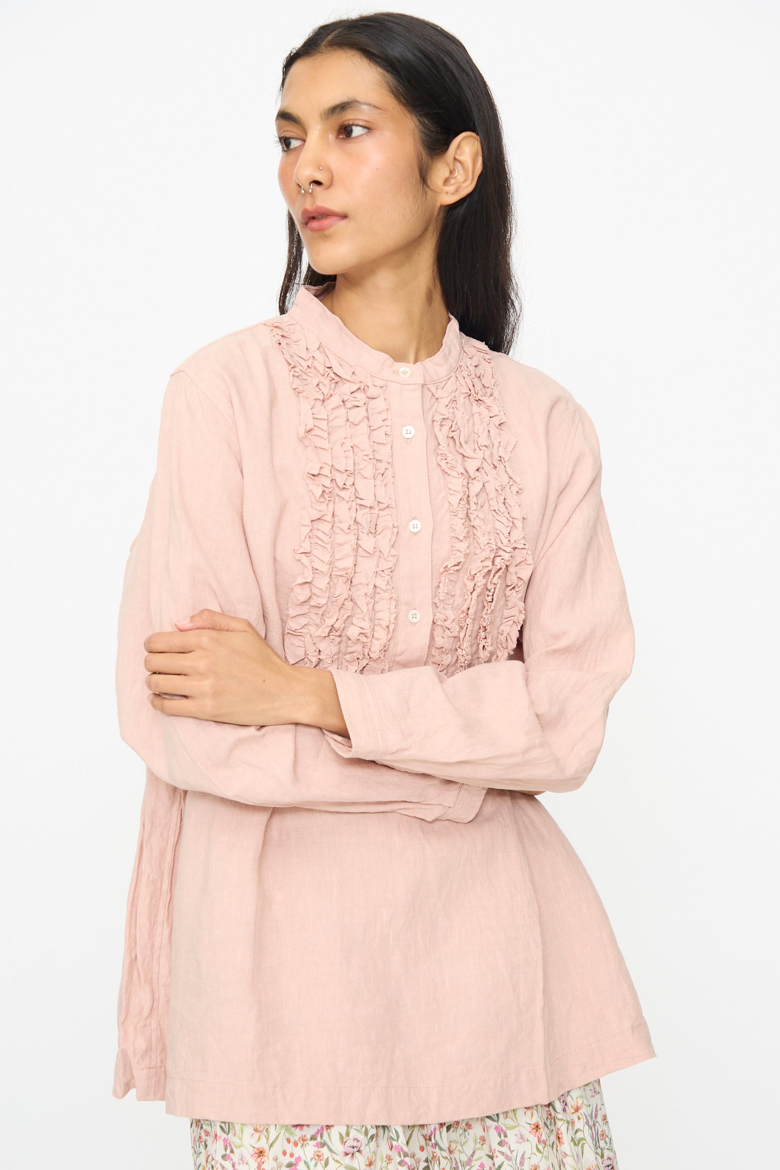 A woman stands with arms crossed, looking to the side, wearing the Ichi Antiquités French Linen Shirt in Pink—a long-sleeve shirt with ruffle detail on the front and a band collar—against a plain background.