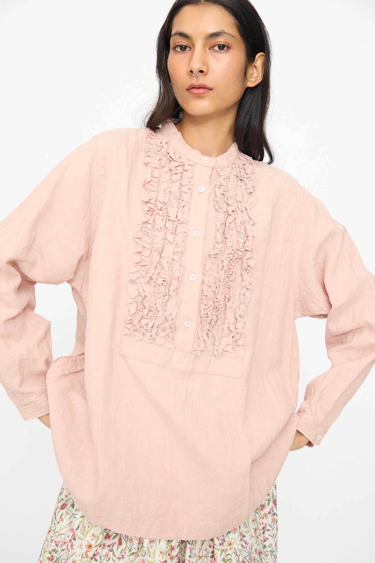 A woman stands against a white background wearing the Ichi Antiquités French Linen Shirt in Pink, featuring a band collar, long sleeves, front ruffle detail, and paired with a floral skirt.