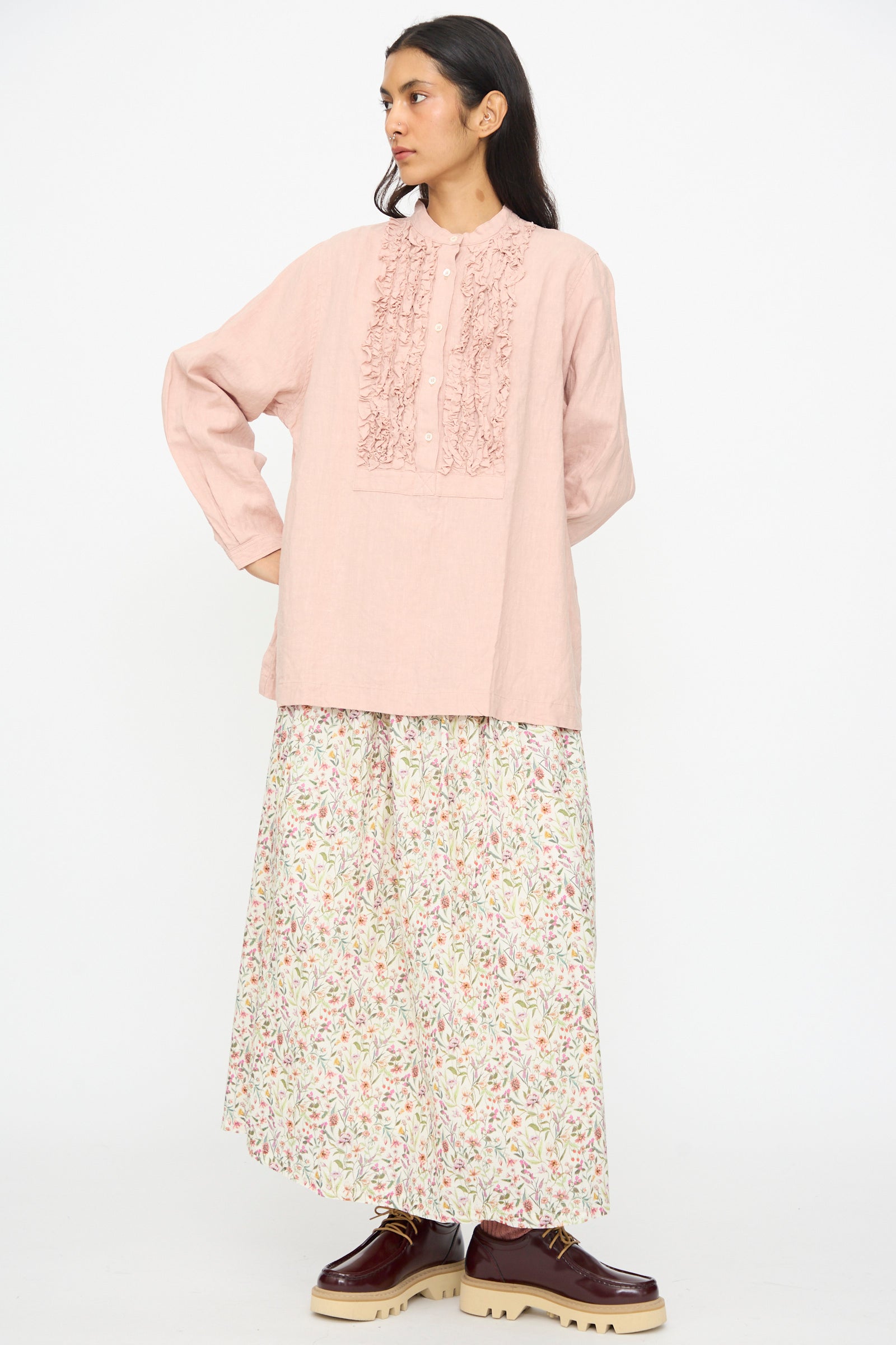 A woman with long dark hair wears the French Linen Shirt in Pink by Ichi Antiquités—featuring a band collar and textured ruffle details—paired with a long, floral-patterned skirt and brown platform shoes against a plain background.