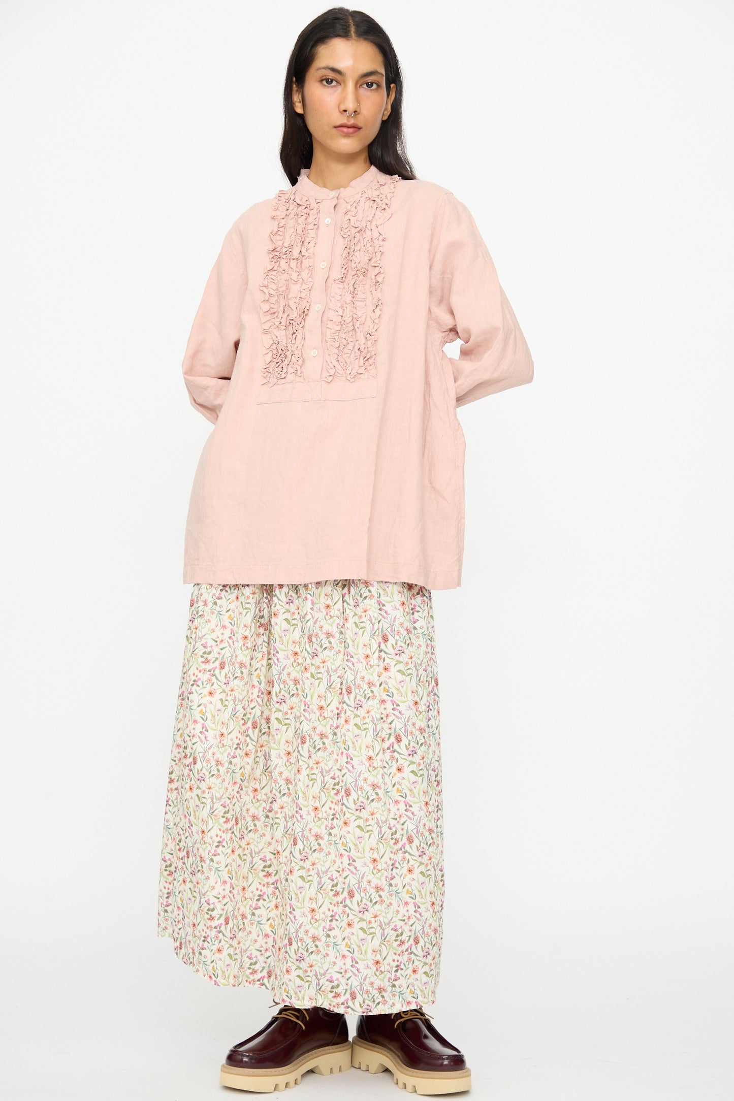 A person wears the Ichi Antiquités French Linen Shirt in Pink, styled with a long floral skirt and brown platform shoes, standing against a plain white background.