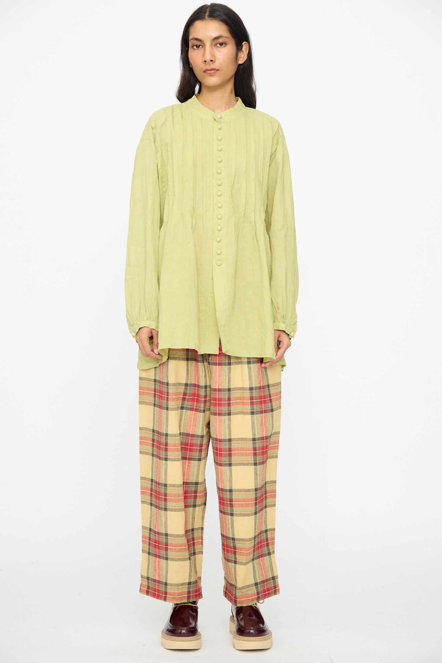Person stands against a white background wearing Ichi Antiquités' French Linen Shirt in Pistachio, paired with loose plaid beige pants with red stripes and brown shoes. This stylish Made in Japan outfit blends comfort and elegance.