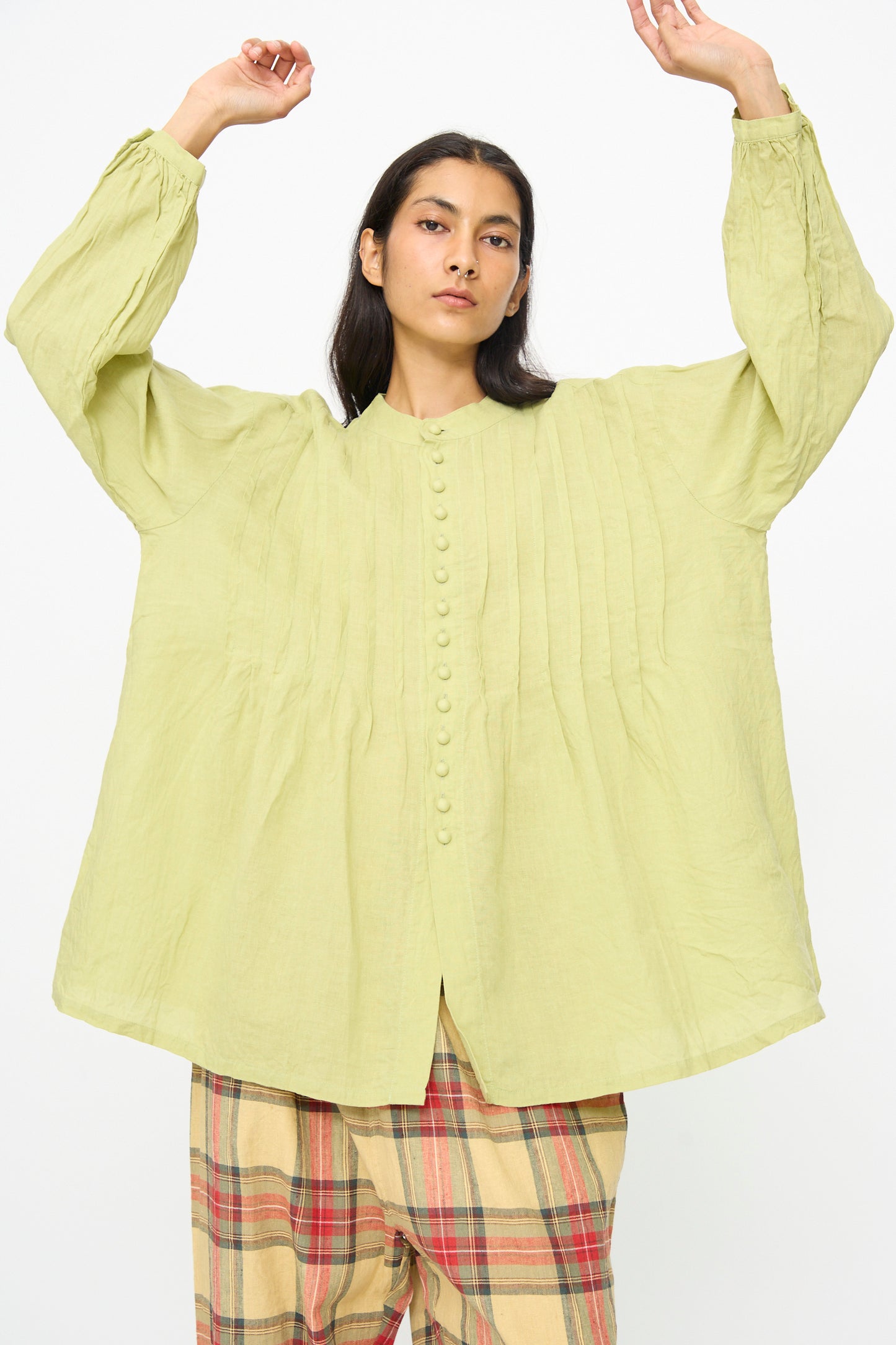 A person wearing the Ichi Antiquités French Linen Shirt in Pistachio, featuring pintuck details and a band collar, paired with plaid pants, stands with arms raised against a plain background.