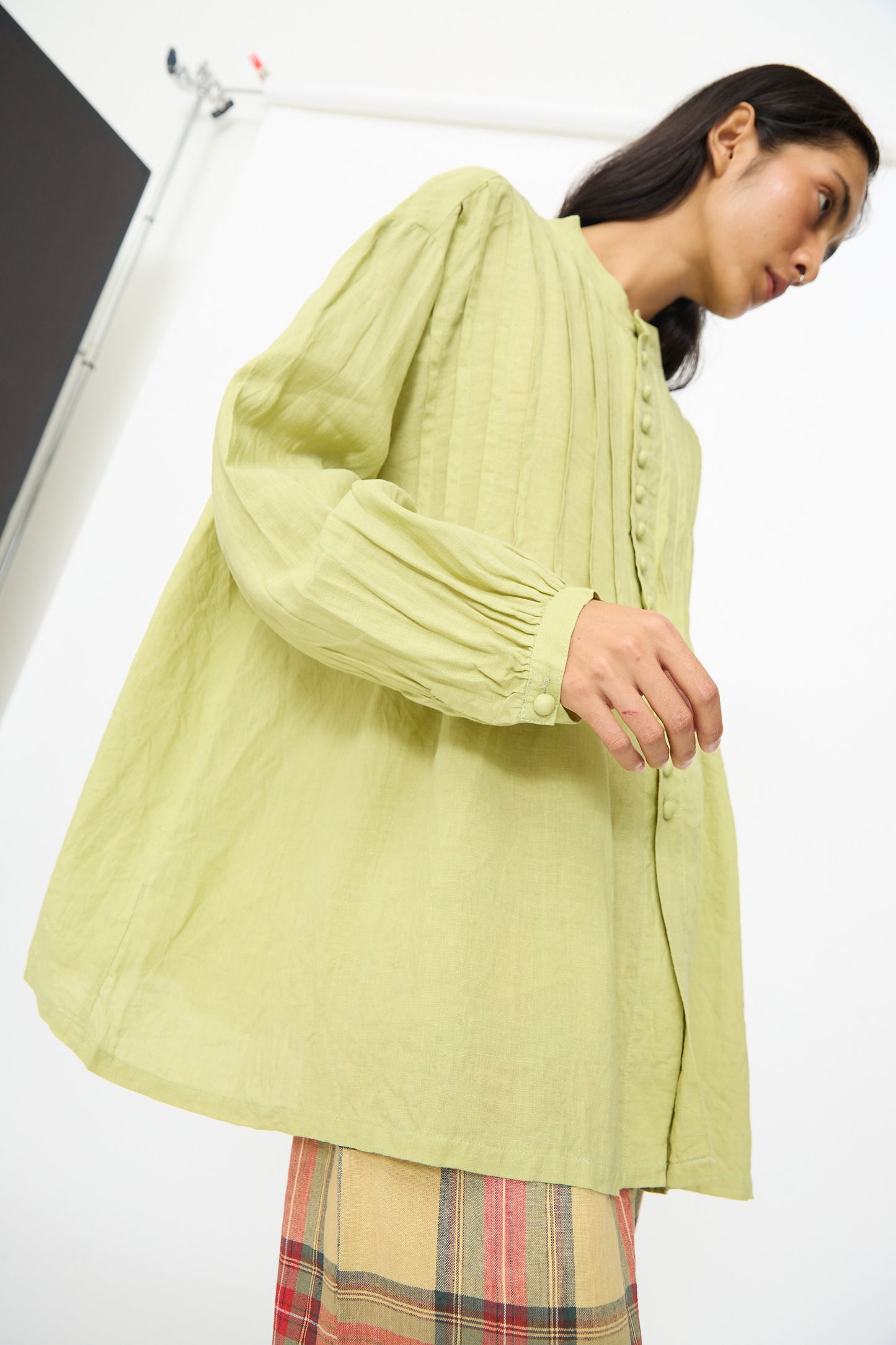 A person wears the Ichi Antiquités French Linen Shirt in Pistachio, featuring a band collar and pleated detailing, paired with a plaid skirt and standing against a plain white background.