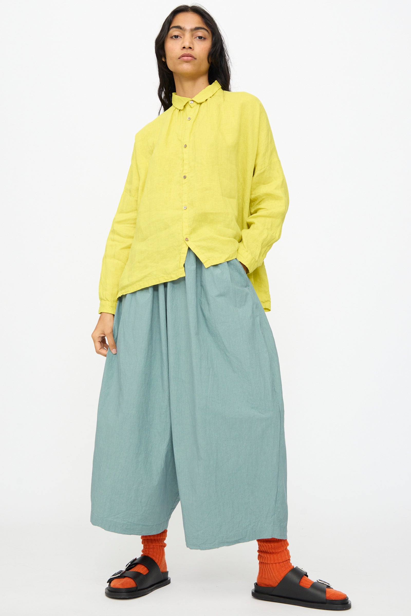 A person stands against a plain white background wearing the Ichi Antiquités French Linen Shirt in Yellow, styled with wide light blue pants, orange socks, and black sandals.