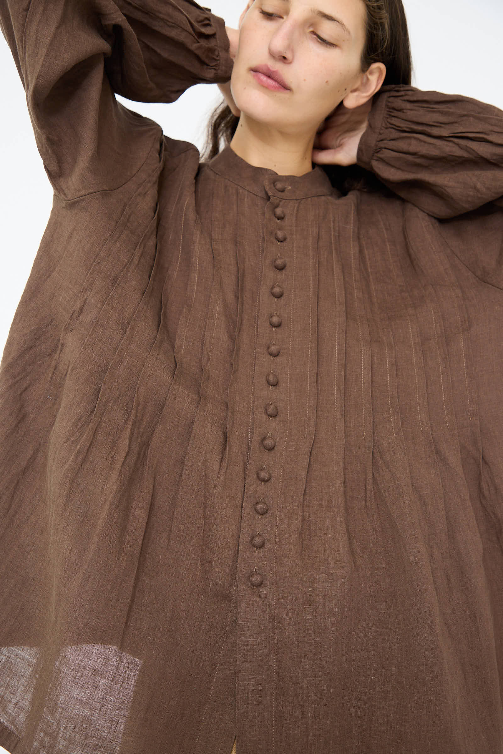 A person wearing the Ichi Antiquités French Linen Shirt in Brown, which features pleated details, a relaxed fit, and a band collar, poses with their hands behind their head.