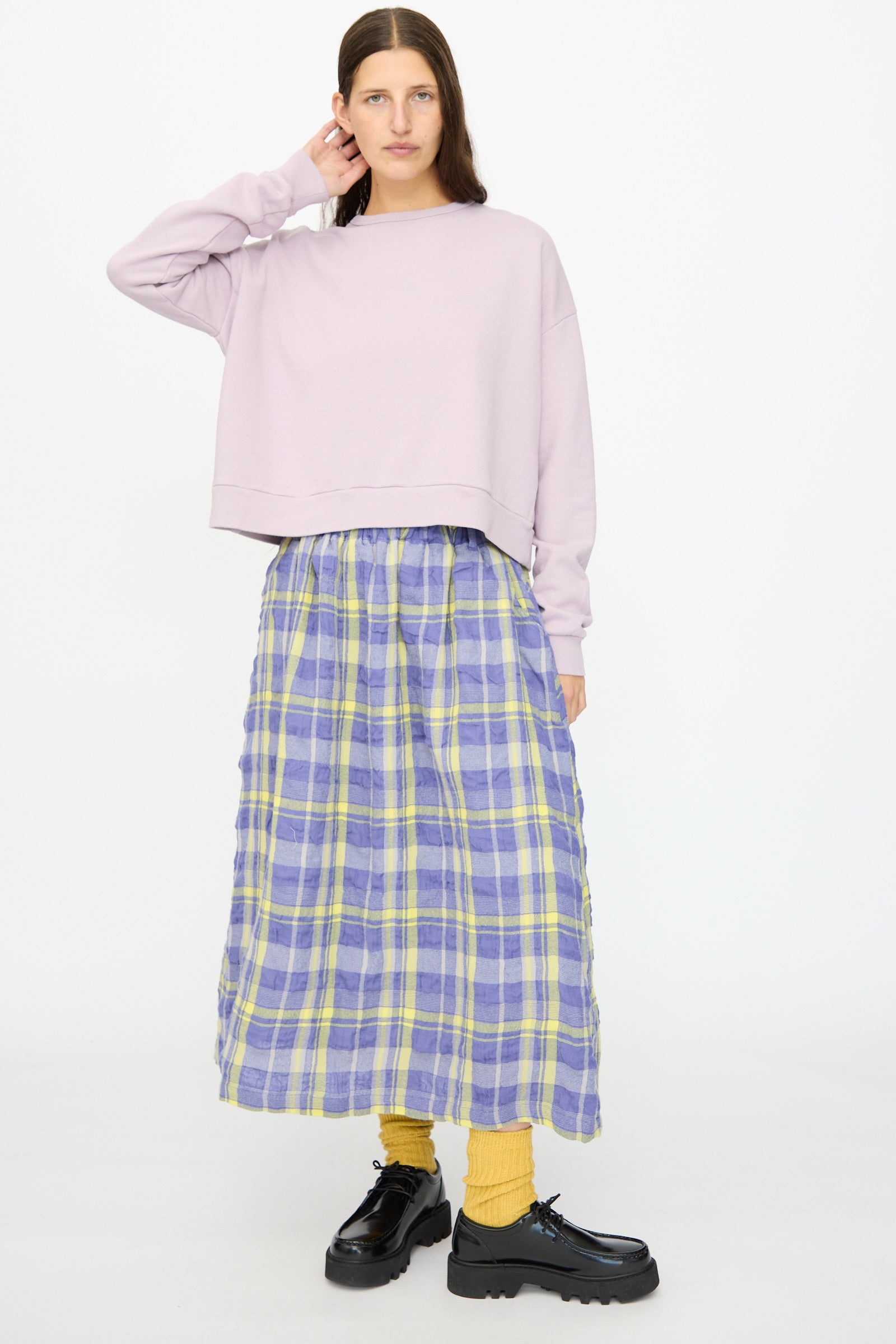 A person stands against a white background wearing the Ichi Antiquités French Terry Cotton Pullover in Lavender, paired with a blue and yellow plaid skirt, yellow socks, and black platform shoes.