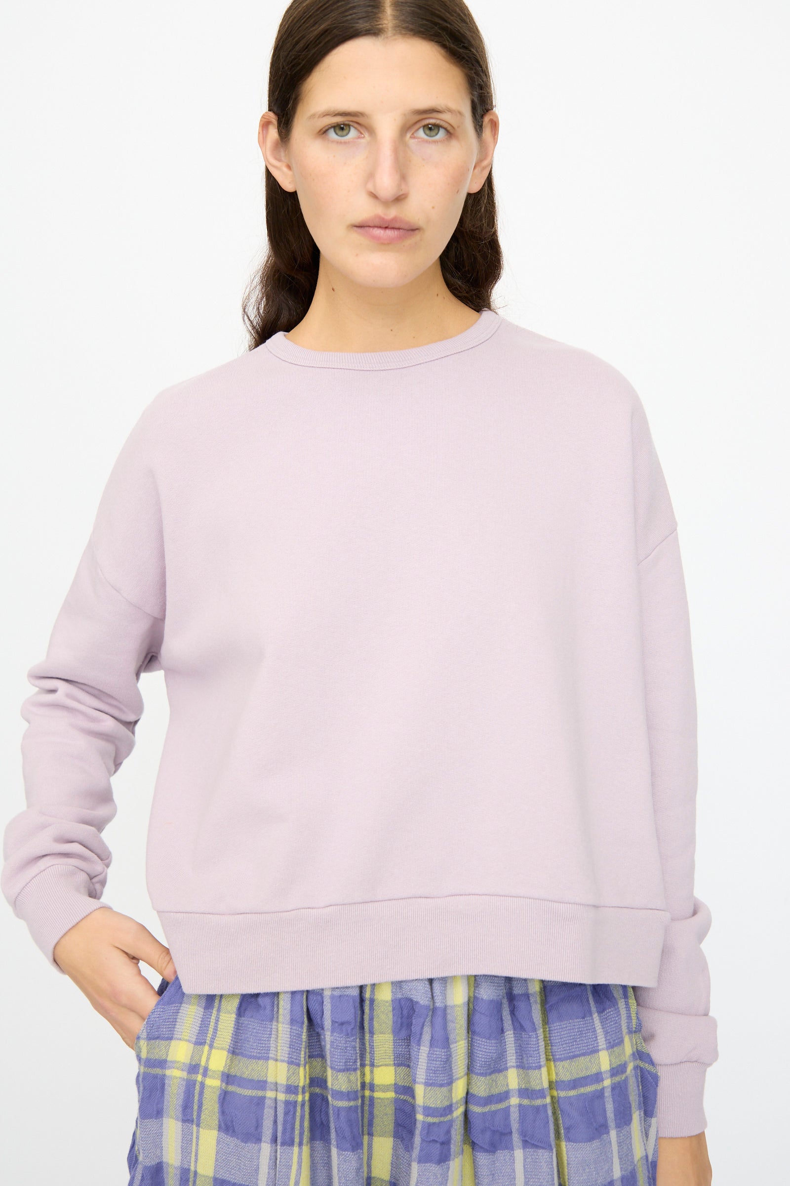 A person with long brown hair models the Ichi Antiquités French Terry Cotton Pullover in Lavender, paired with a blue and yellow plaid skirt, against a plain white background.