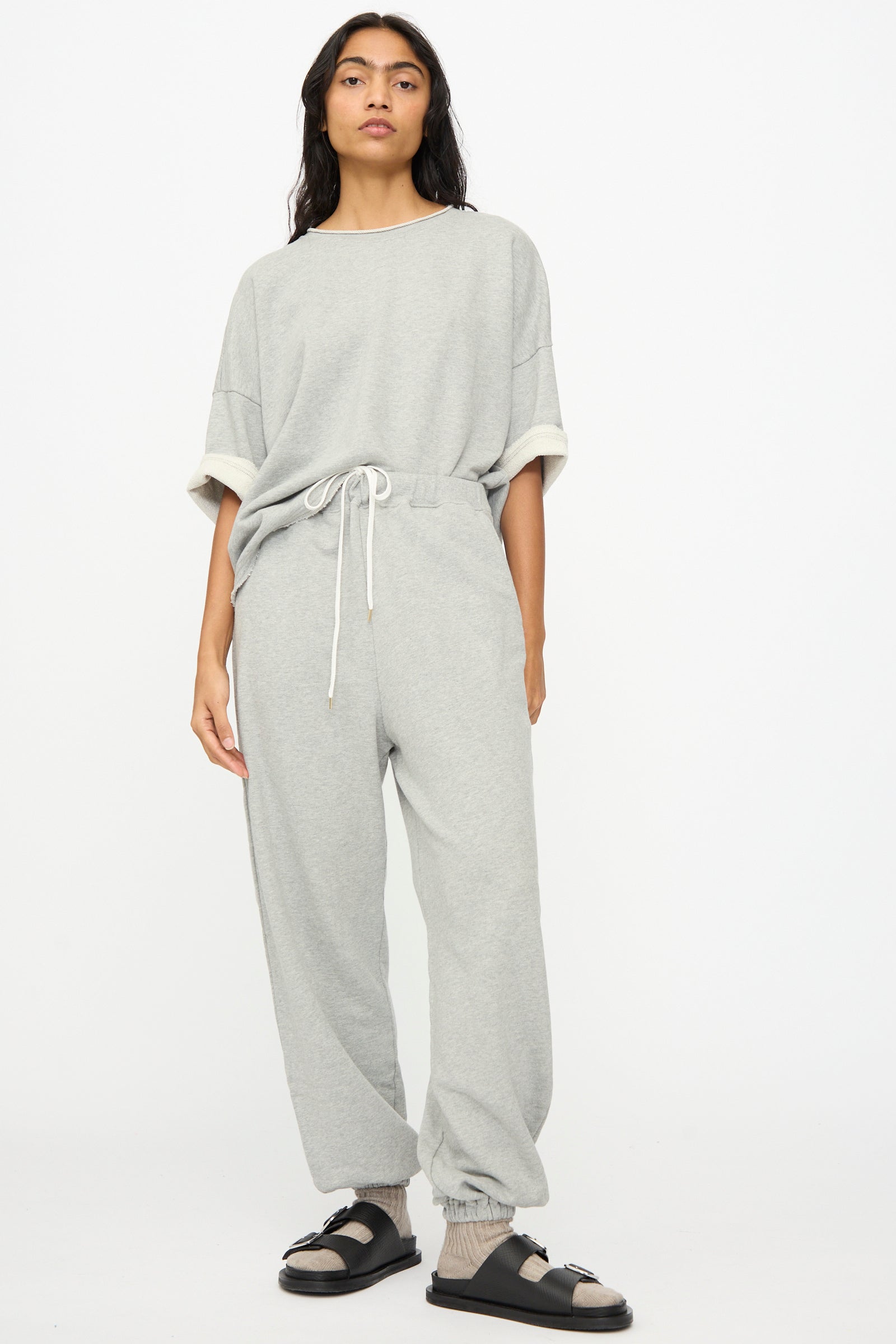 A person stands against a white background wearing Ichi Antiquités French Terry Pants in Gray, a loose grey sweatshirt, and black sandals with socks.