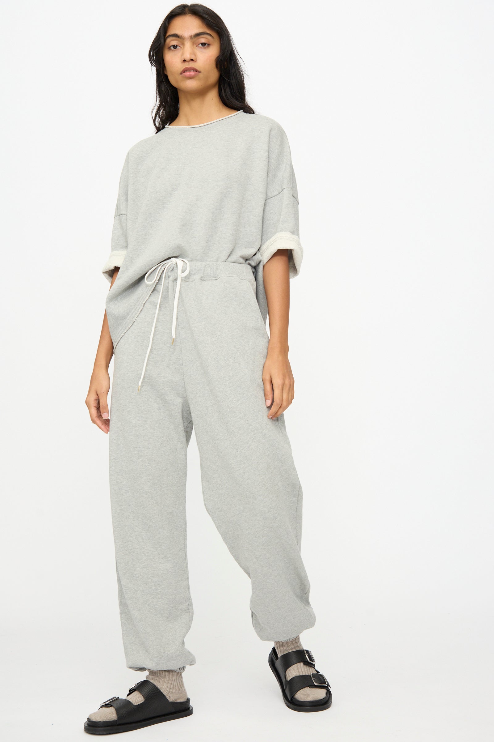 A person faces forward in a loose light gray sweatshirt and Ichi Antiquités French Terry Pants in Gray with a drawstring, complemented by gray socks and black sandals. The background is plain white.