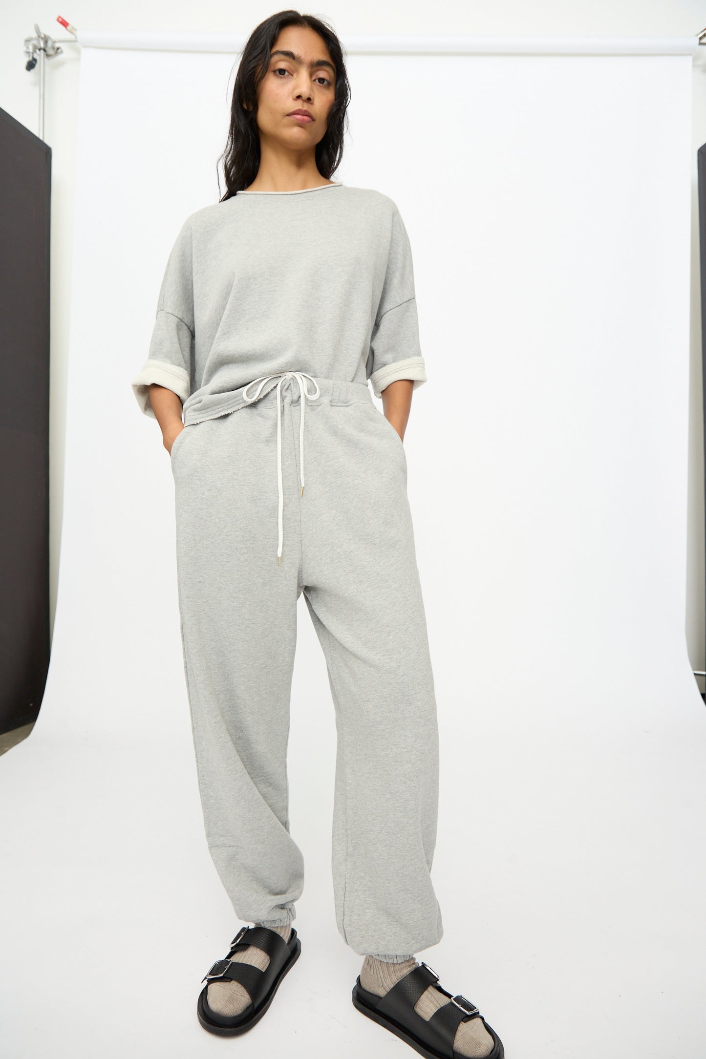 A person stands against a white backdrop wearing Ichi Antiquités French Terry Pants in Gray and a matching top, with hands in pockets, black sandals, and beige socks.