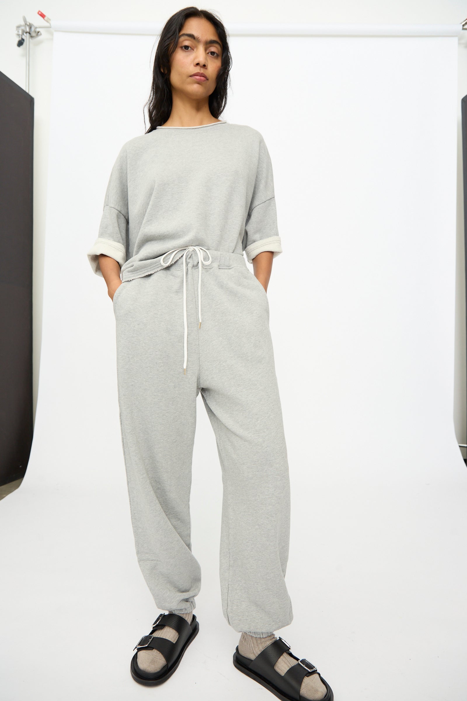 A person stands against a white backdrop wearing Ichi Antiquités French Terry Pants in Gray and a matching top, with hands in pockets, black sandals, and beige socks.