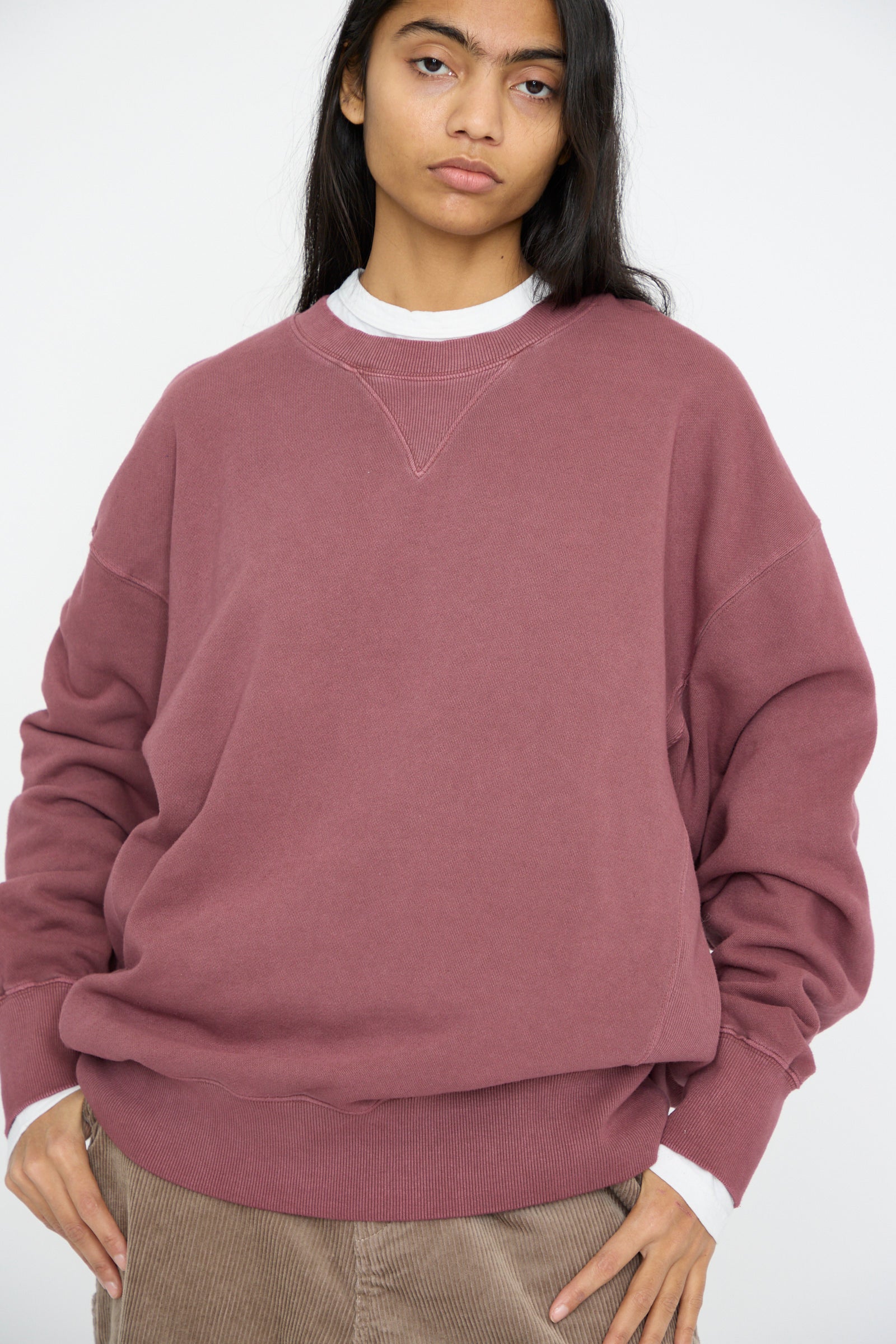 A person wearing an Ichi Antiquités French Terry Pigment Pullover in Burgundy, teamed with brown pants, stands with their hands in pockets against a simple background.