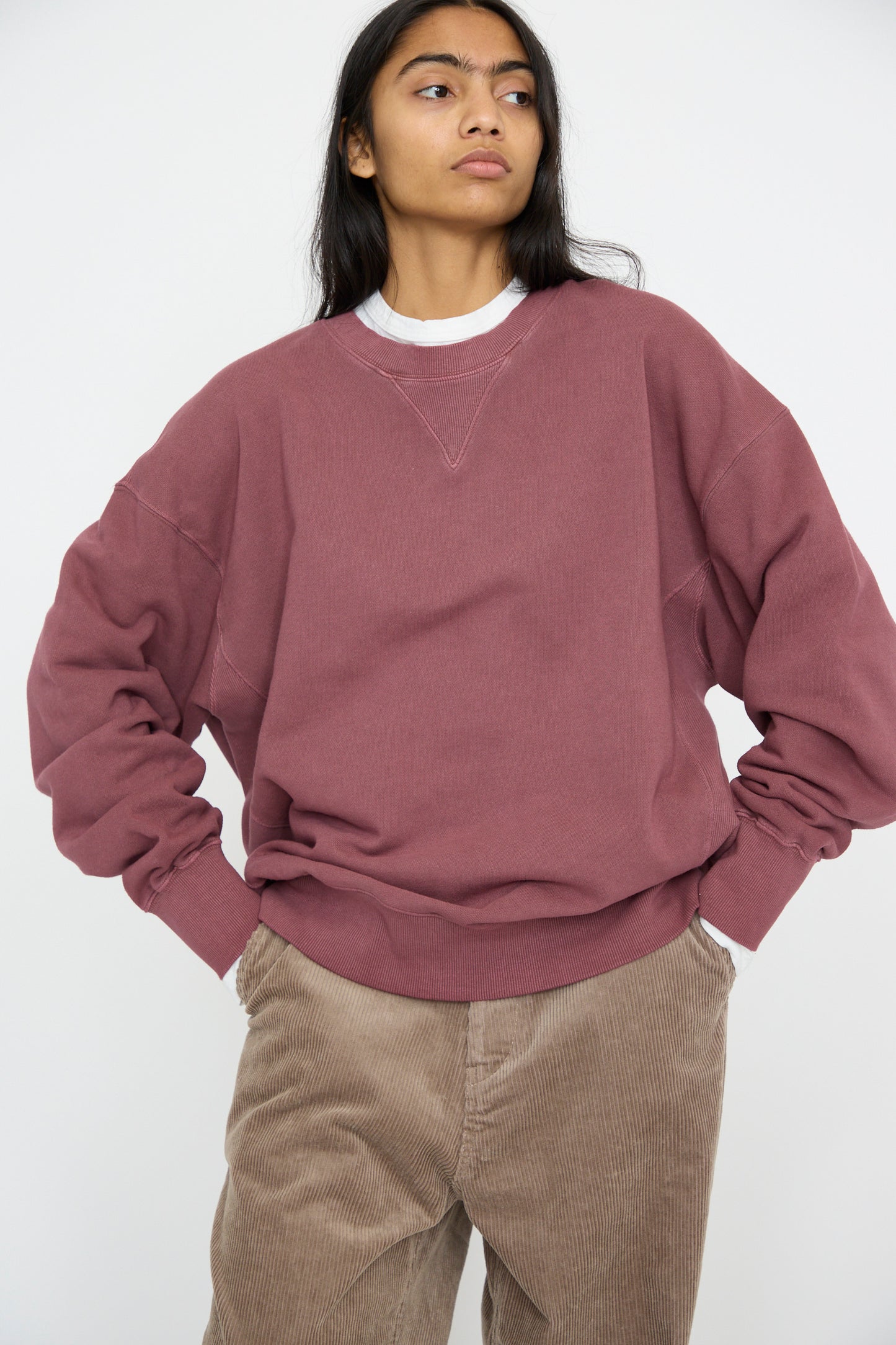 A person with long hair wearing an Ichi Antiquités French Terry Pigment Pullover in Burgundy and brown corduroy pants stands with hands on hips against a plain background.