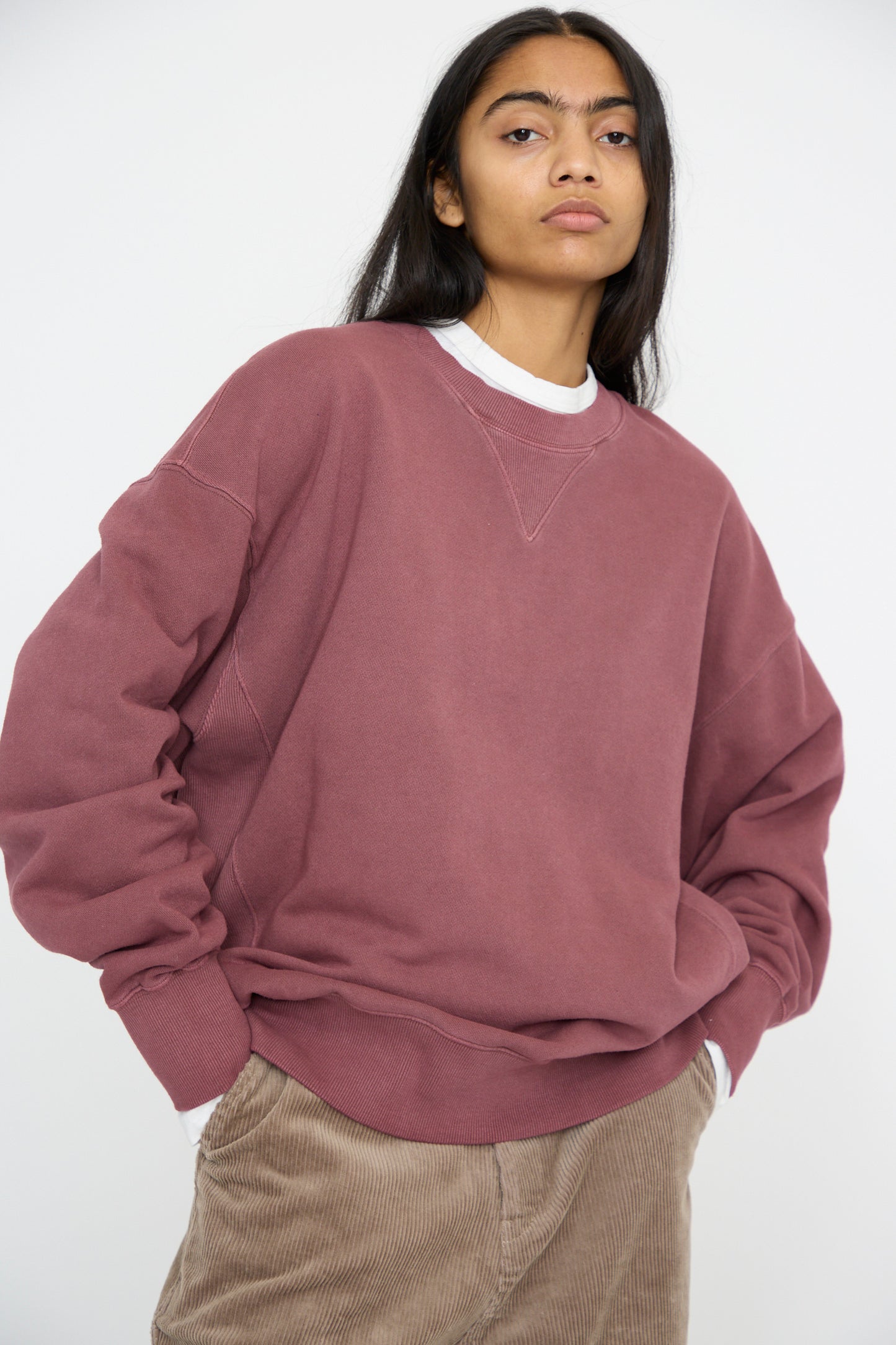 A person is standing against a plain background, wearing an oversized French Terry Pigment Pullover in Burgundy by Ichi Antiquités over a white shirt and brown corduroy pants.