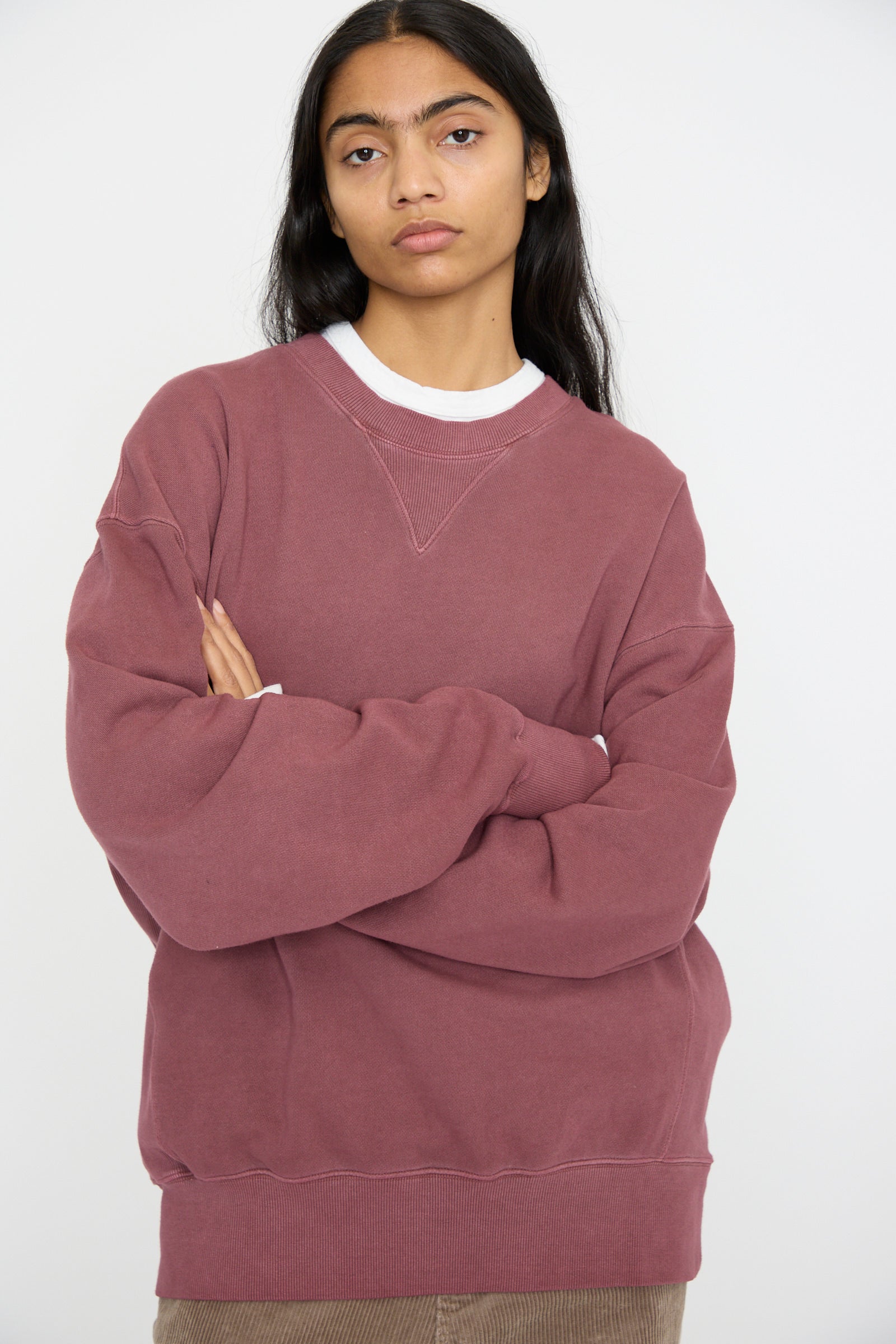 A person stands against a white background, wearing the French Terry Pigment Pullover in Burgundy by Ichi Antiquités and brown pants, arms crossed.