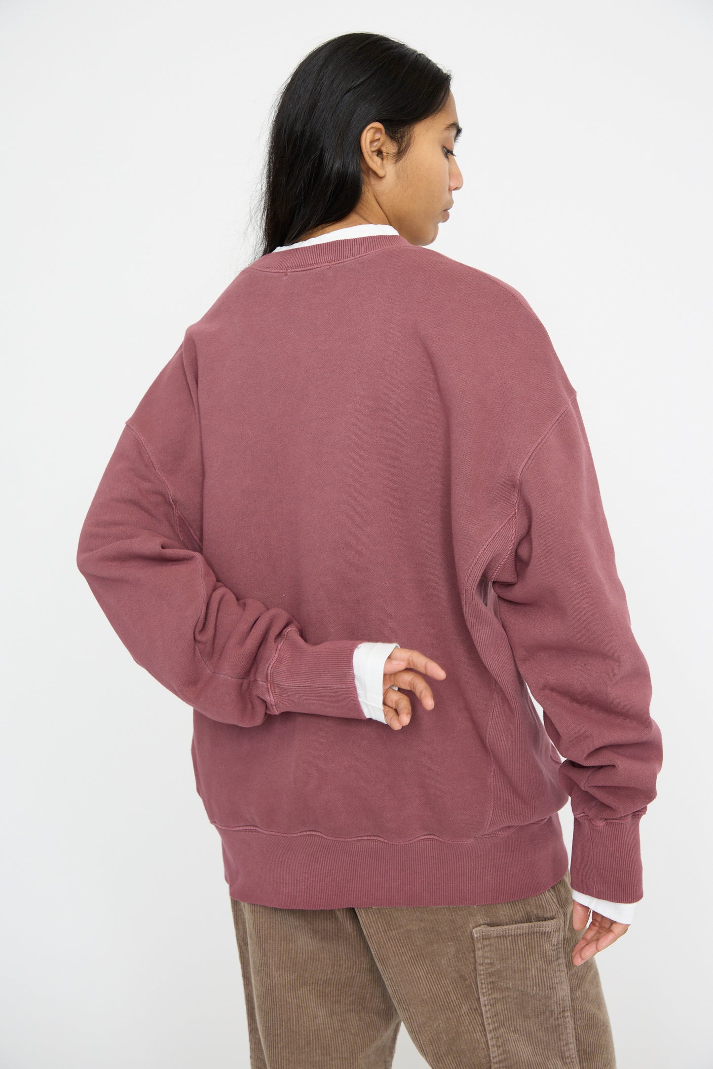 A person in an oversized French Terry Pigment Pullover in Burgundy by Ichi Antiquités stands with their back to the camera, one arm behind their back.