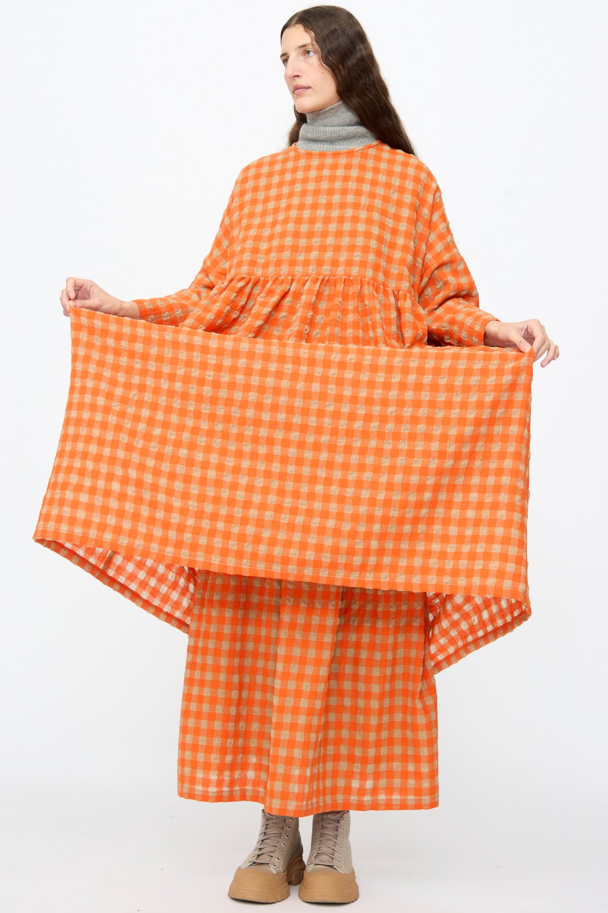 A person wearing an Ichi Antiquités Gingham Check Dress in Beige and Orange over a grey mock-neck top and beige boots, lifting the skirt to show the wool cotton blend fabric.