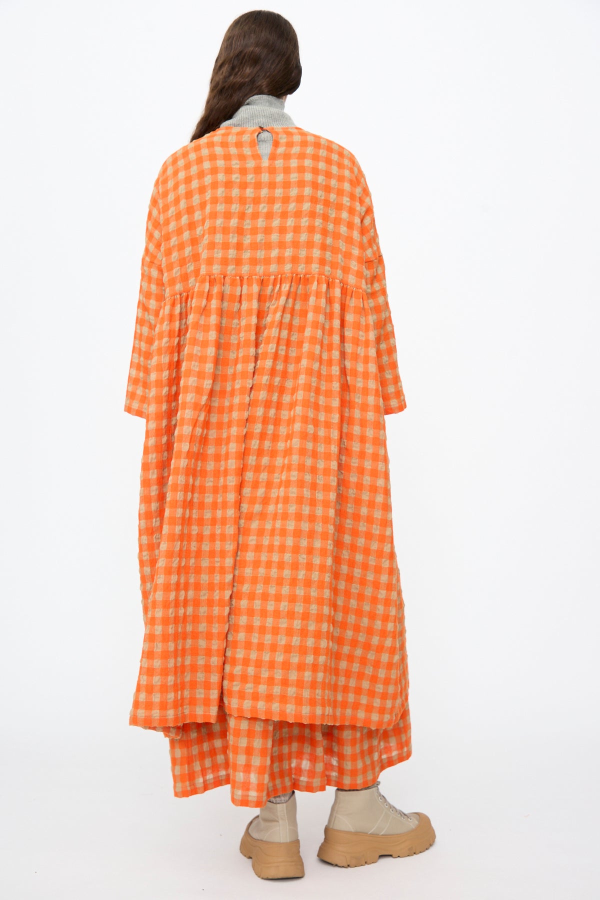 Seen from behind, a person wears an Ichi Antiquités Gingham Check Dress in Beige and Orange, featuring long sleeves, a dropped waist, and a long skirt, paired with beige lace-up boots.