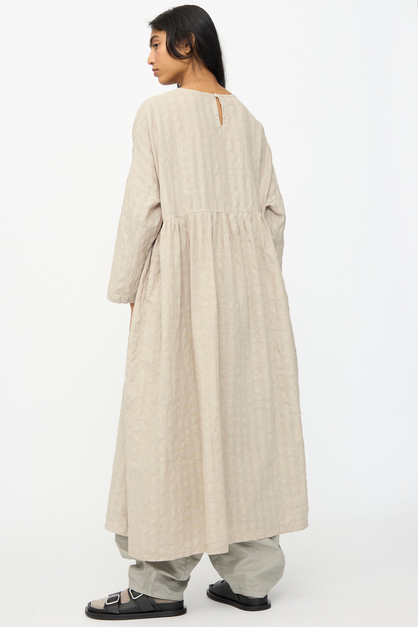 A person stands sideways against a plain white background, wearing the Ichi Antiquités Gingham Check Dress in Natural, styled with wide-leg pants and sandals.