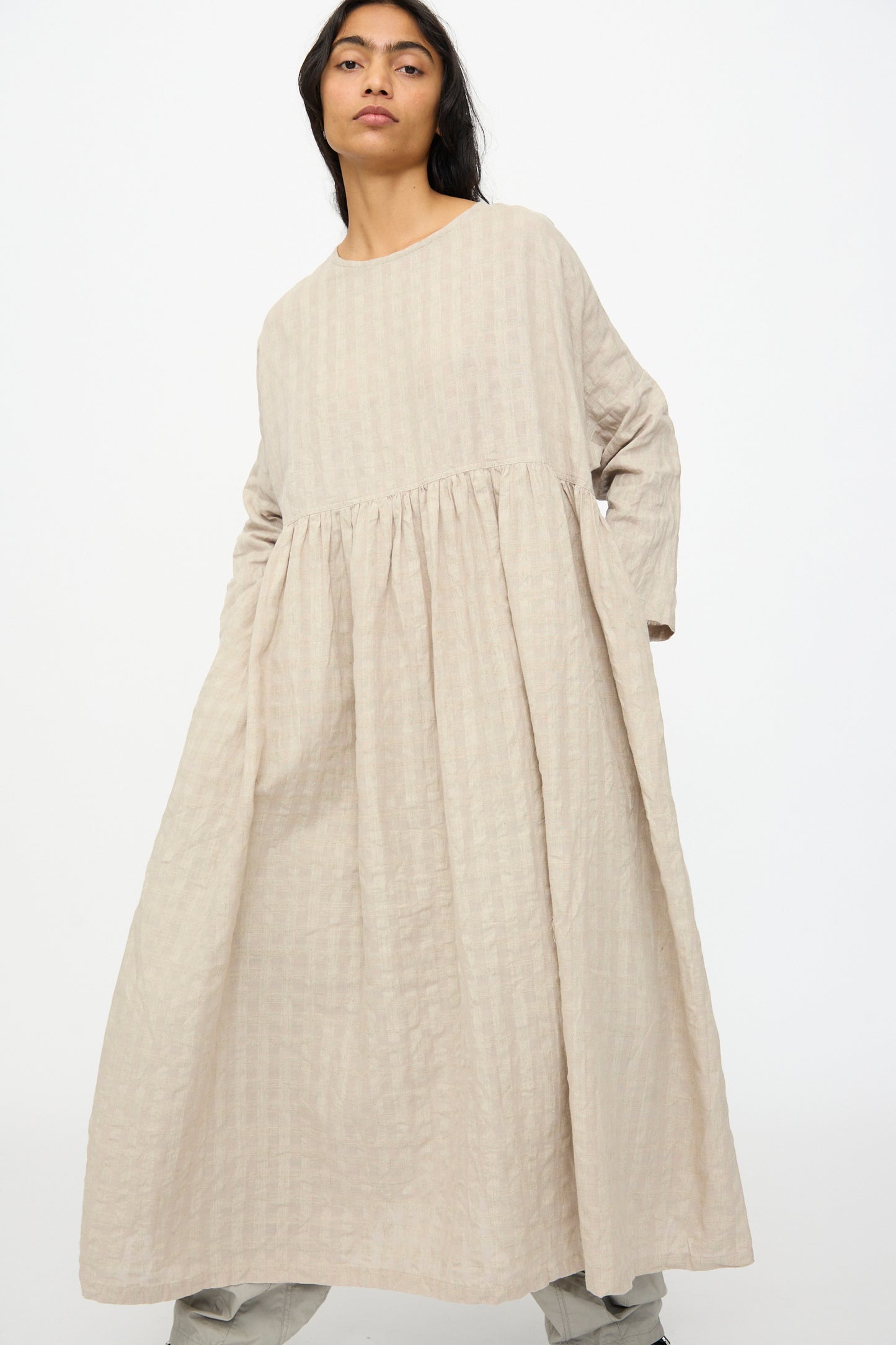 A person wearing the Ichi Antiquités Gingham Check Dress in Natural, a loose-fitting long sleeve piece made from a cotton linen blend, stands against a plain white background.