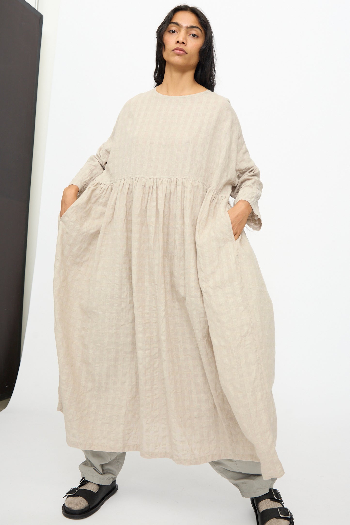 A person stands against a plain background wearing the Ichi Antiquités Gingham Check Dress in Natural, paired with grey pants and black sandals.