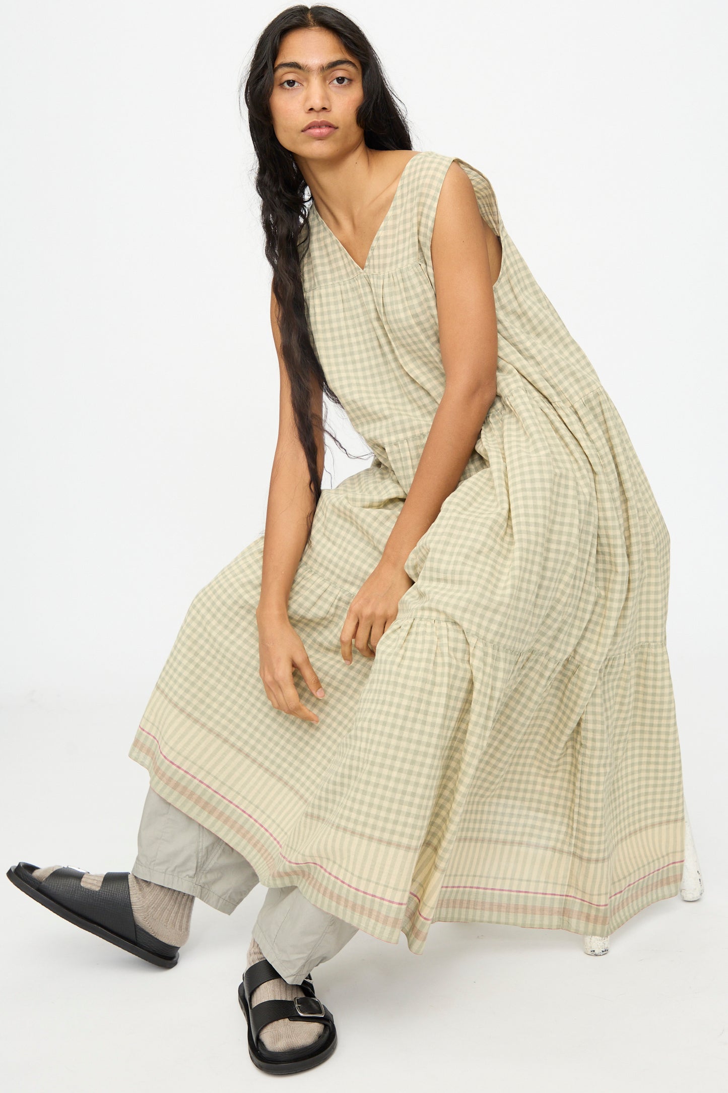 A person with long dark hair wears the Ichi Antiquités Gingham Check Khadi Cotton Dress in Khaki over light pants and black sandals, seated against a plain white background.