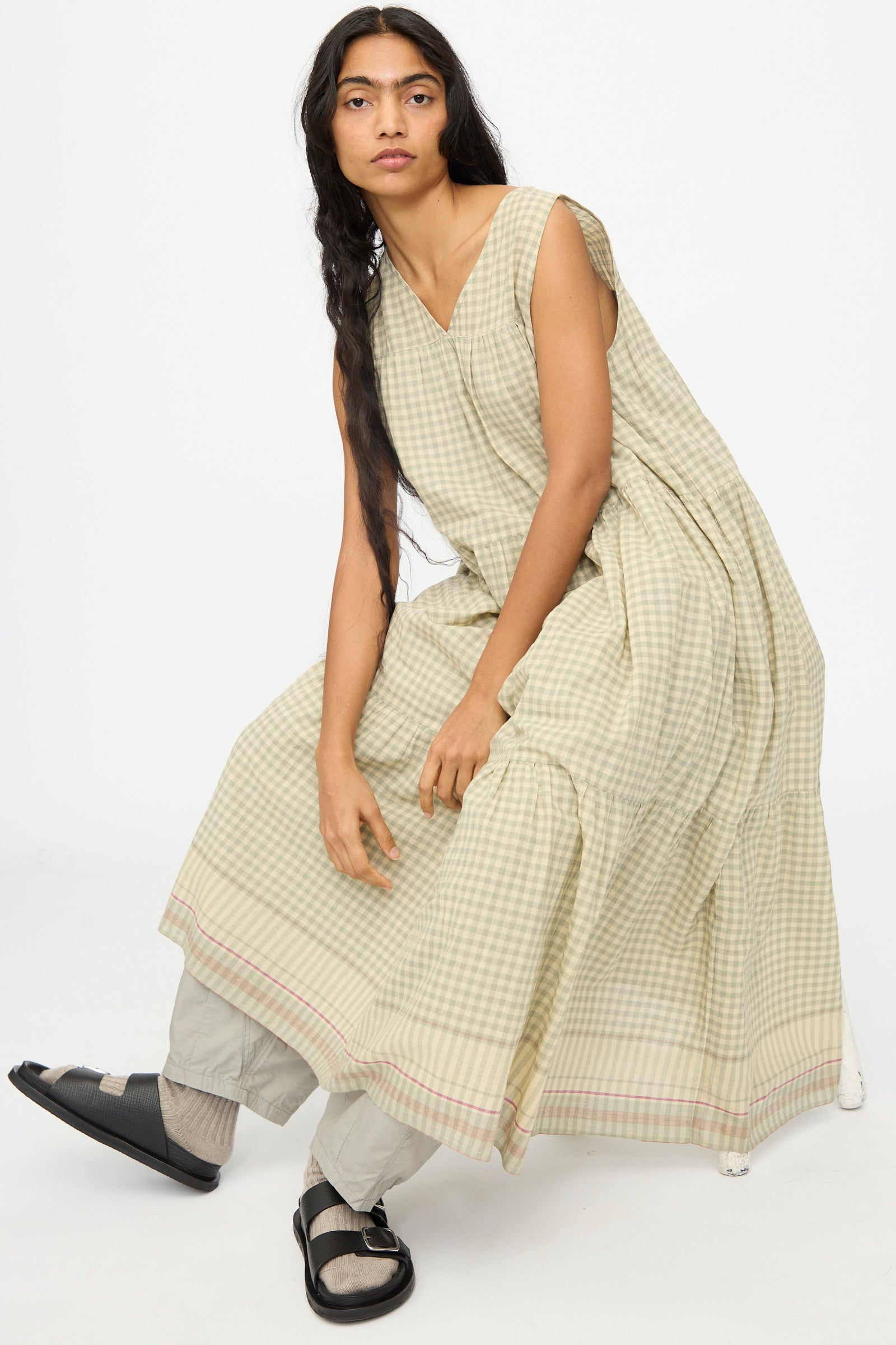A person with long dark hair wears the Ichi Antiquités Gingham Check Khadi Cotton Dress in Khaki over light pants and black sandals, seated against a plain white background.