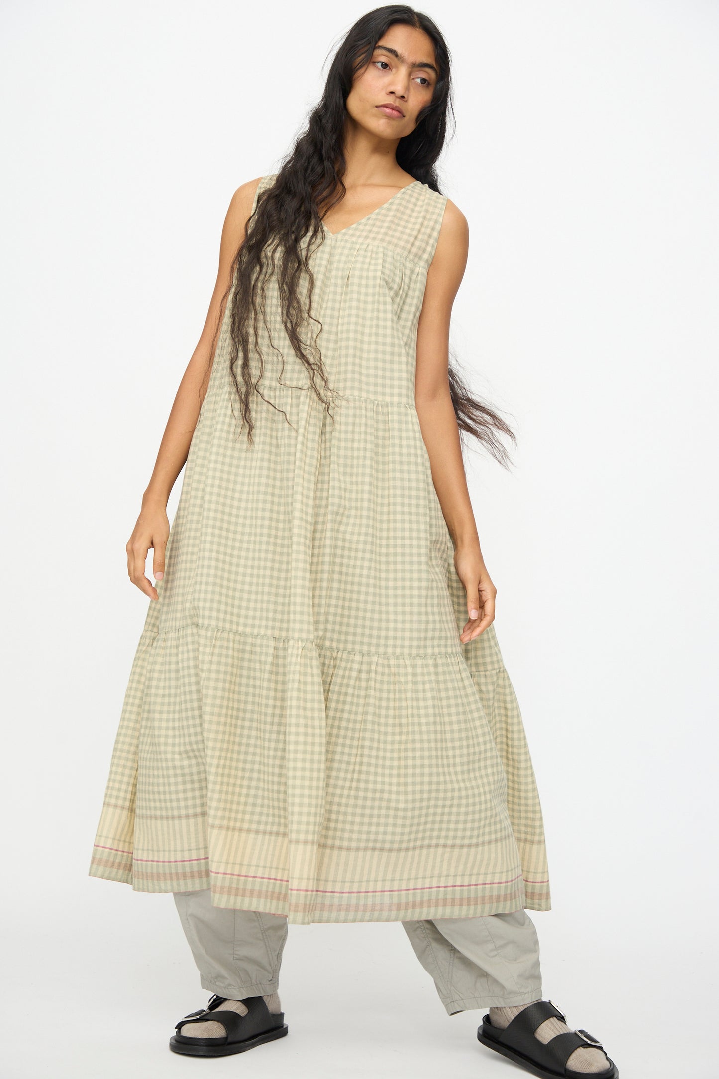 A person with long hair wears the Ichi Antiquités Gingham Check Khadi Cotton Dress in khaki over gray pants and black sandals, standing against a plain white background.