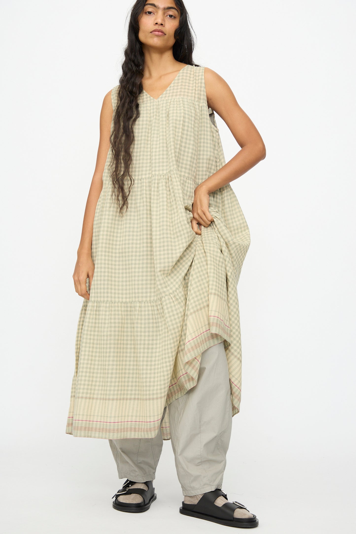 A person wears the Ichi Antiquités Gingham Check Khadi Cotton Dress in khaki over light gray pants with black sandals, standing against a plain white background.