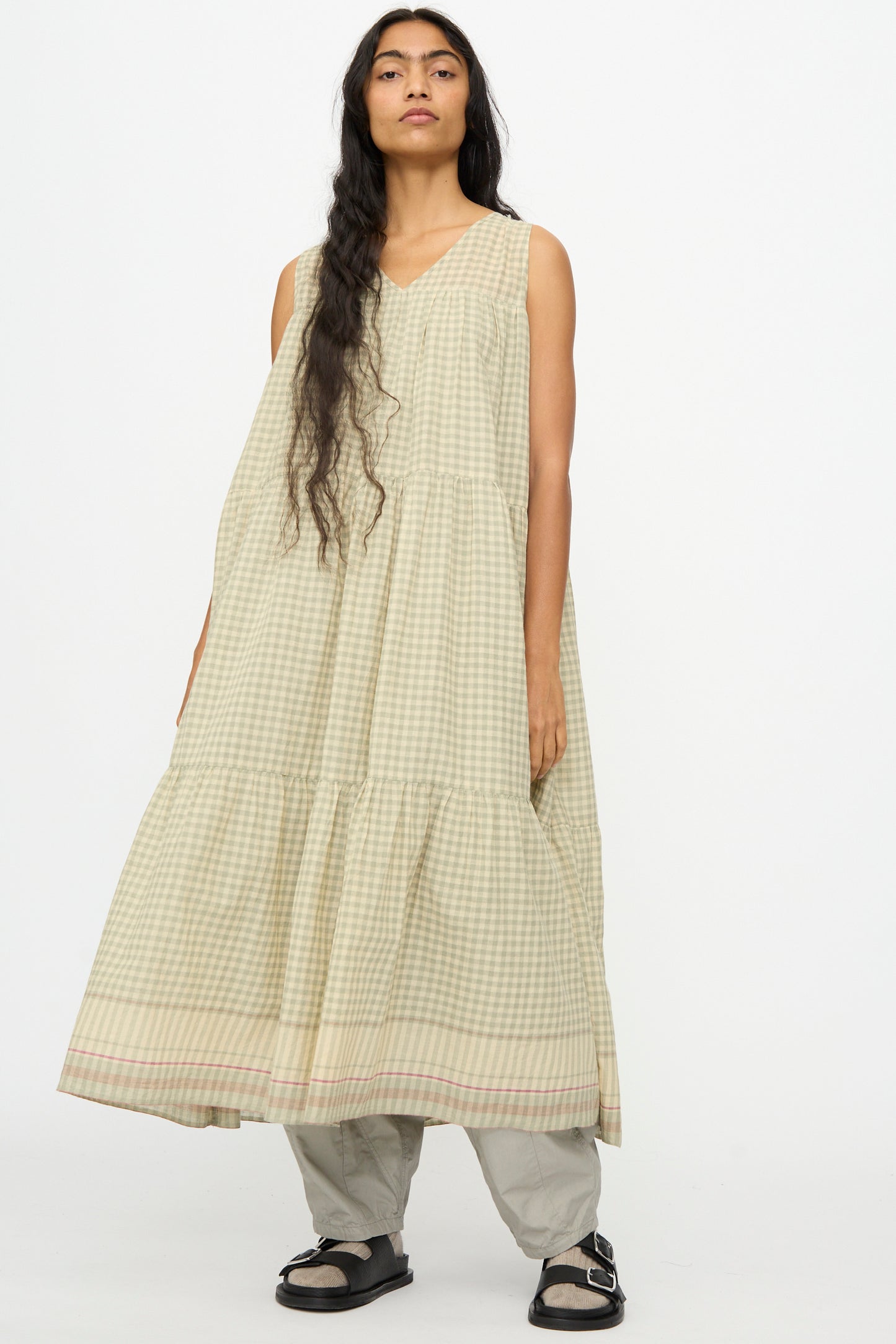 A person with long hair wears the Ichi Antiquités Gingham Check Khadi Cotton Dress in Khaki over wide-legged pants and sandals, standing against a plain white background.