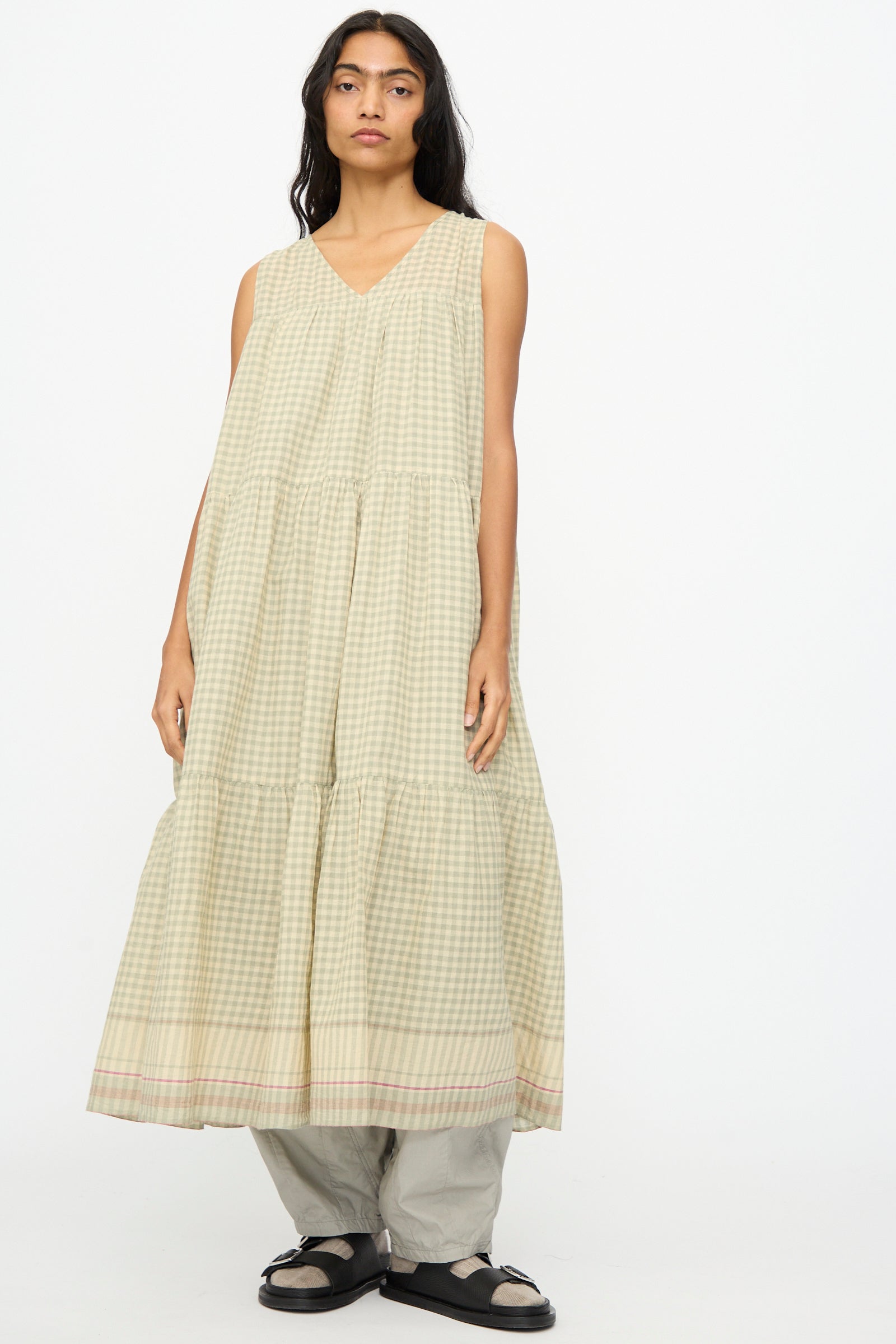 A person wearing the Ichi Antiquités Gingham Check Khadi Cotton Dress in Khaki over light gray pants and black sandals, standing against a plain white background.