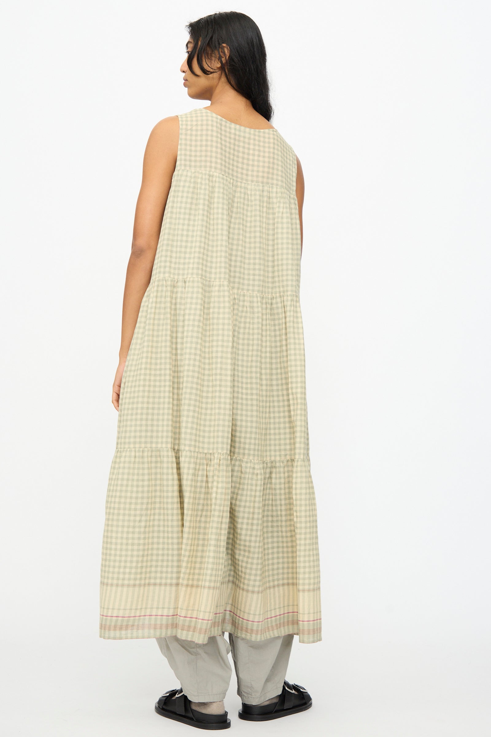 A person stands facing away in the Ichi Antiquités Gingham Check Khadi Cotton Dress in Khaki, layered over light pants with black sandals, set against a plain white background.