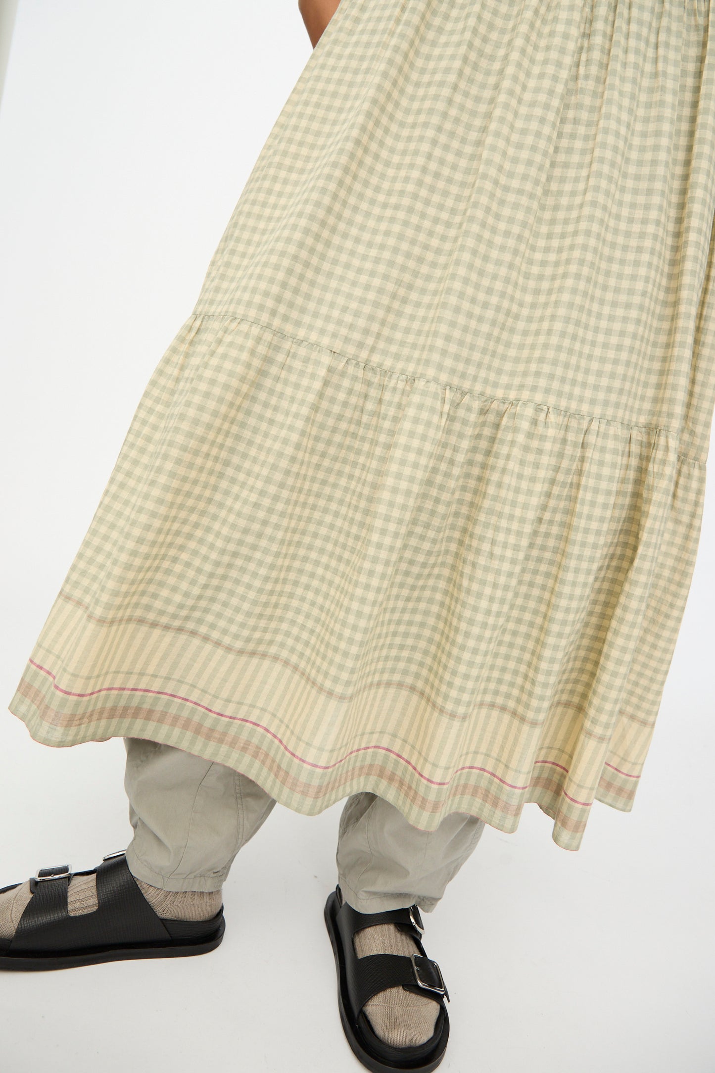 A person wears the Ichi Antiquités Gingham Check Khadi Cotton Dress in Khaki over beige pants, paired with gray socks and black sandals, standing against a plain white background.