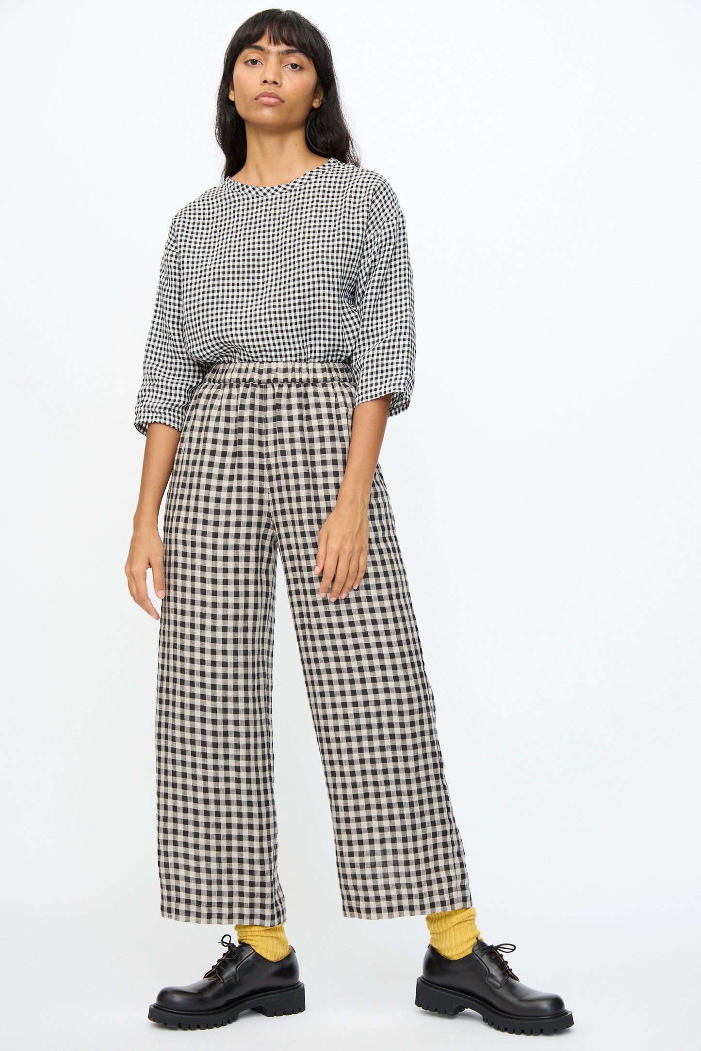 A person stands against a plain light background wearing Ichi Antiquités' Gingham Check Pants in Natural and Black with an elastic waistband, styled with a black-and-white checkered top, yellow socks, and black shoes.