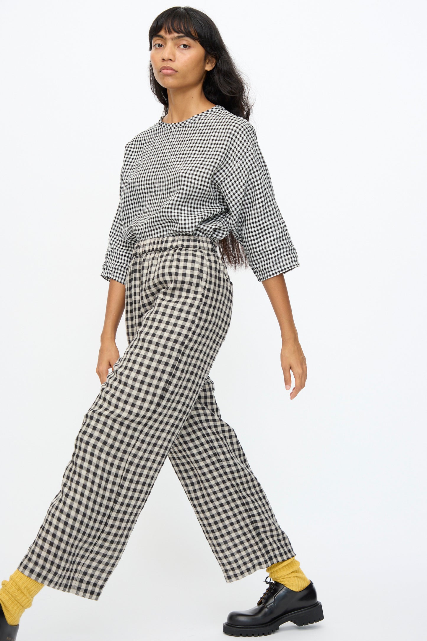 A woman walks against a plain white background wearing Ichi Antiquités Gingham Check Pants in Natural and Black, a black-and-white checkered top, yellow socks, and black shoes.