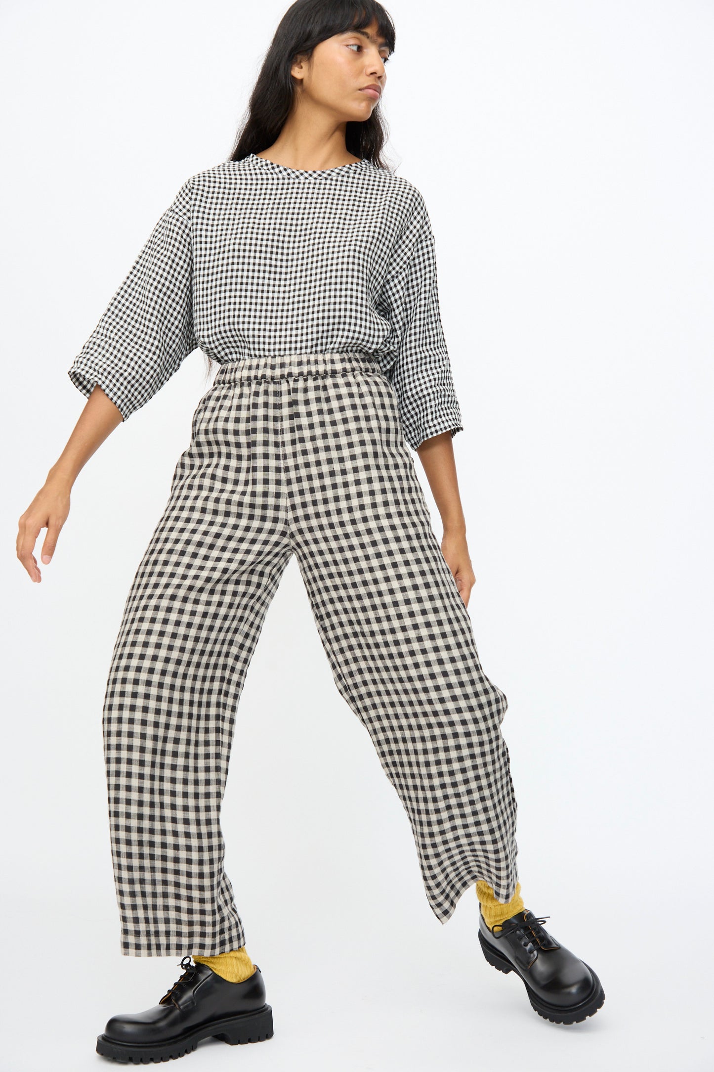 A person wearing Ichi Antiquités Gingham Check Pants in Natural and Black with an elastic waistband stands against a plain white background, paired with yellow socks and black shoes.