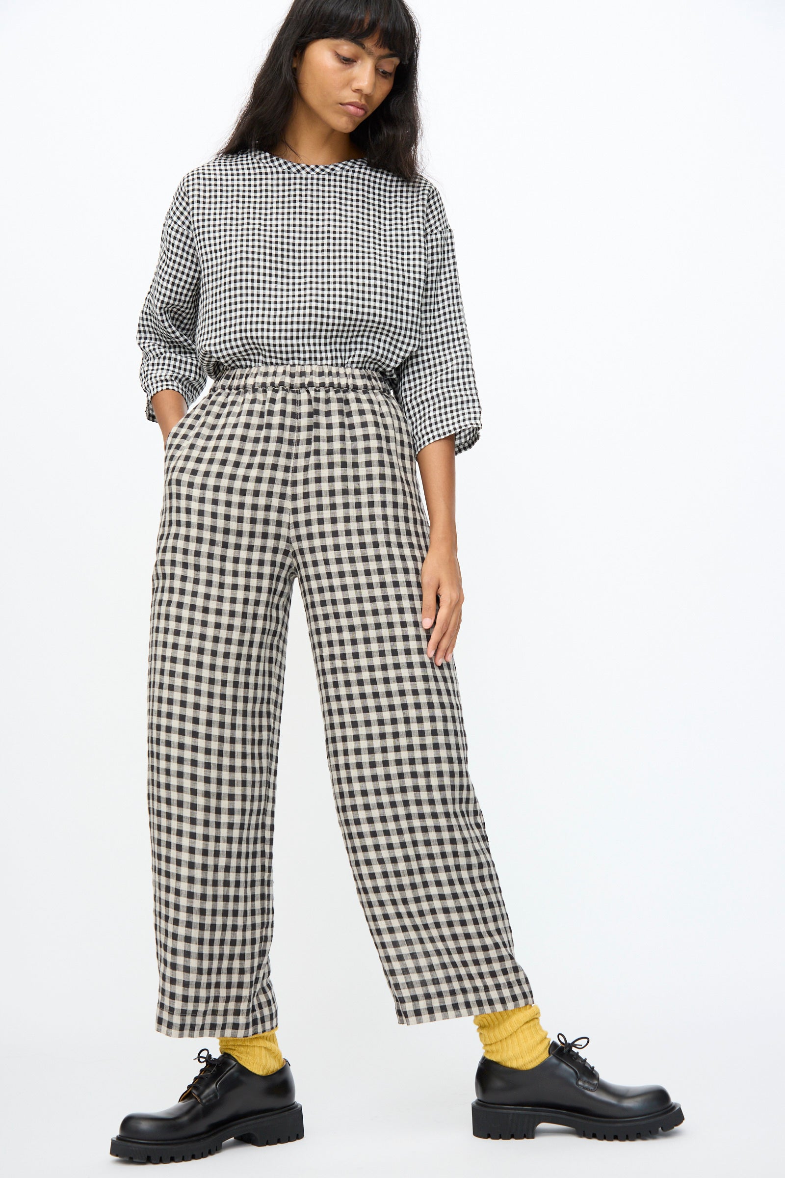 A woman stands against a plain background wearing Ichi Antiquités Gingham Check Pants in Natural and Black, made in Japan with an elastic waistband, paired with a matching top, yellow socks, and black lace-up shoes.
