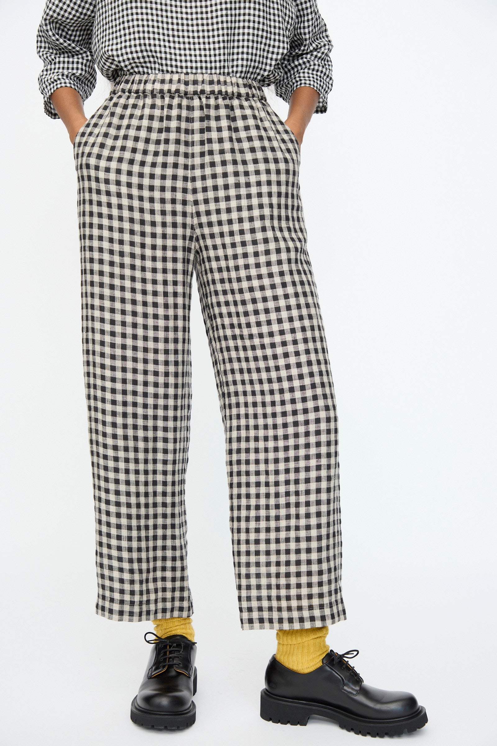A person wears Ichi Antiquités Gingham Check Pants in Natural and Black—Japanese-made linen pants with an elastic waistband—paired with a matching top, yellow socks, and black lace-up shoes, hands in pockets against a plain background.