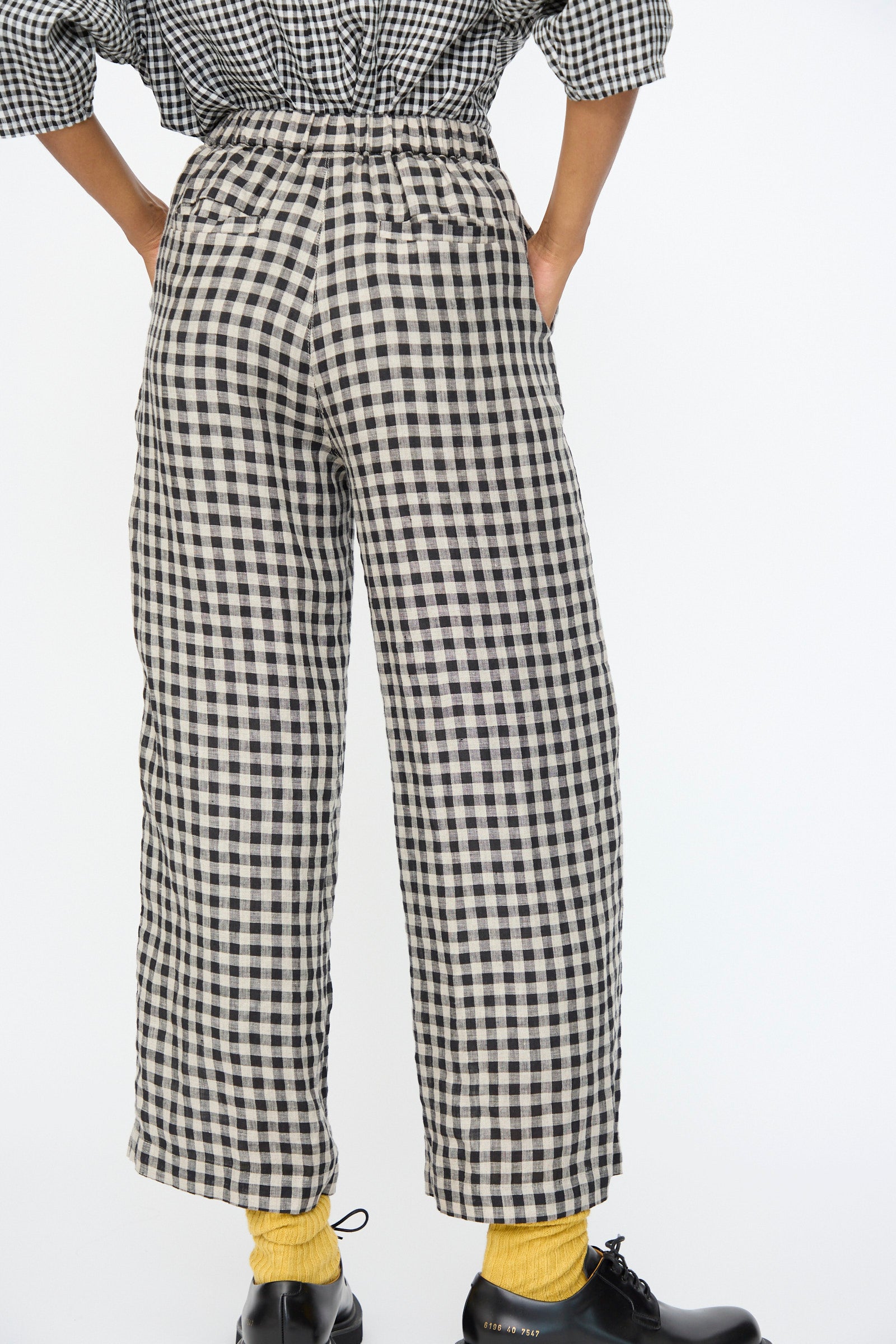 A person wears Ichi Antiquités Gingham Check Pants in Natural and Black—Japanese-made linen with an elastic waistband—paired with a matching top, yellow socks, and black shoes, shown from the back against a plain background.