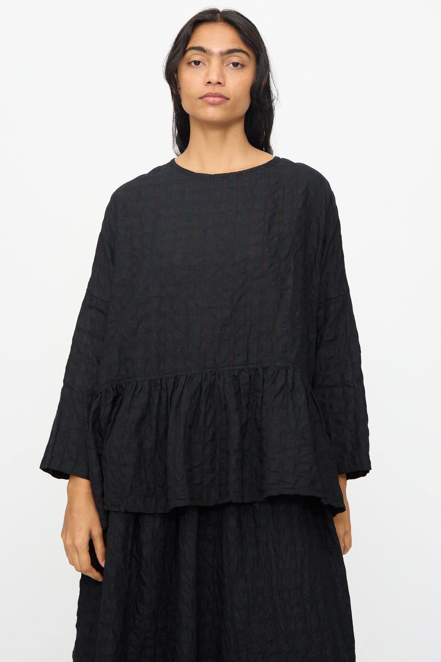 A person wearing the Ichi Antiquités Gingham Check Pullover in Black, a loose-fitting, long-sleeved blouse with a ruffled hem made in Japan, stands against a plain white background.