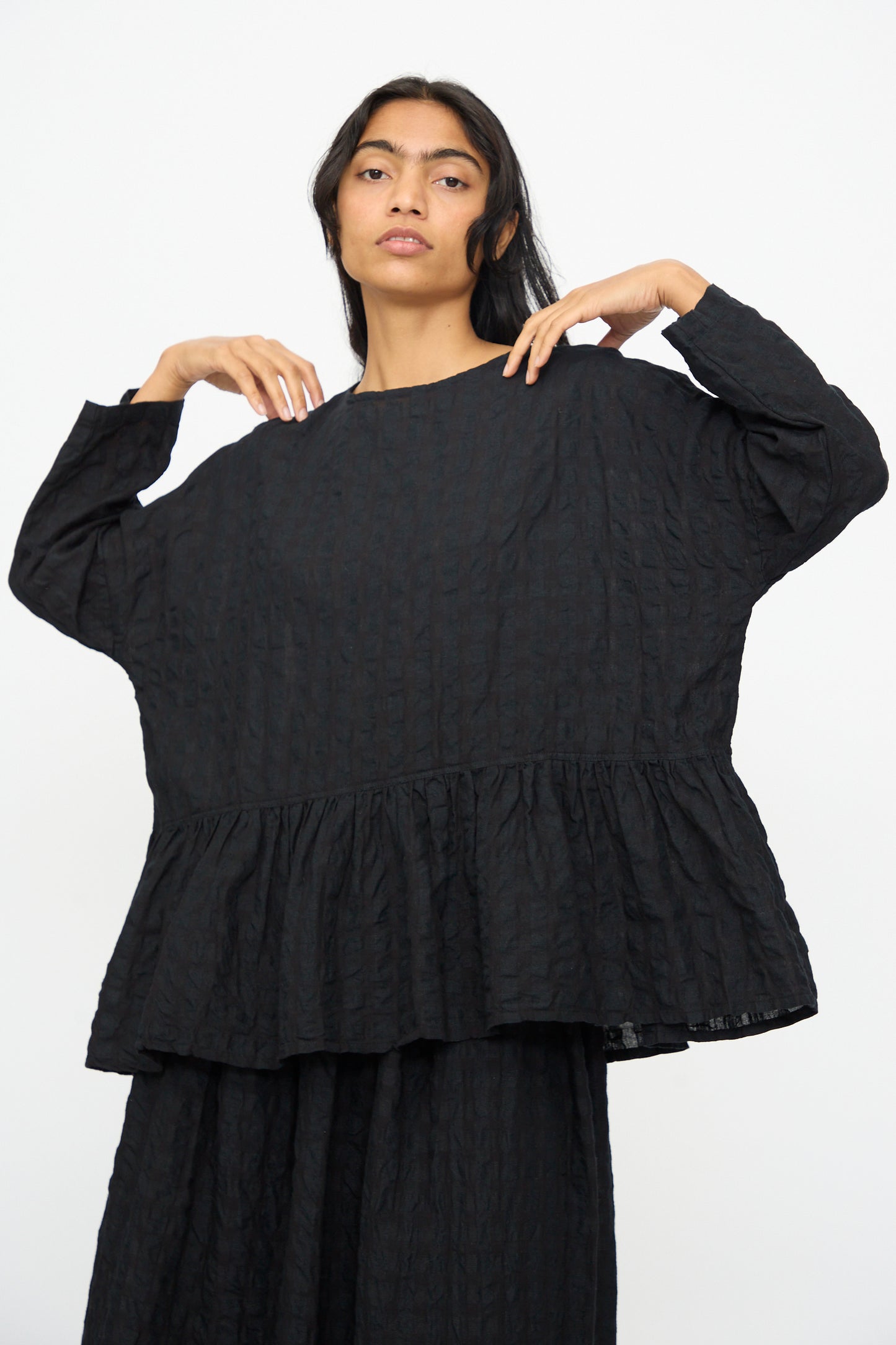 A person models the Ichi Antiquités Gingham Check Pullover in Black, a relaxed fit blouse with a cotton-linen blend and ruffled hem, standing against a plain white background.