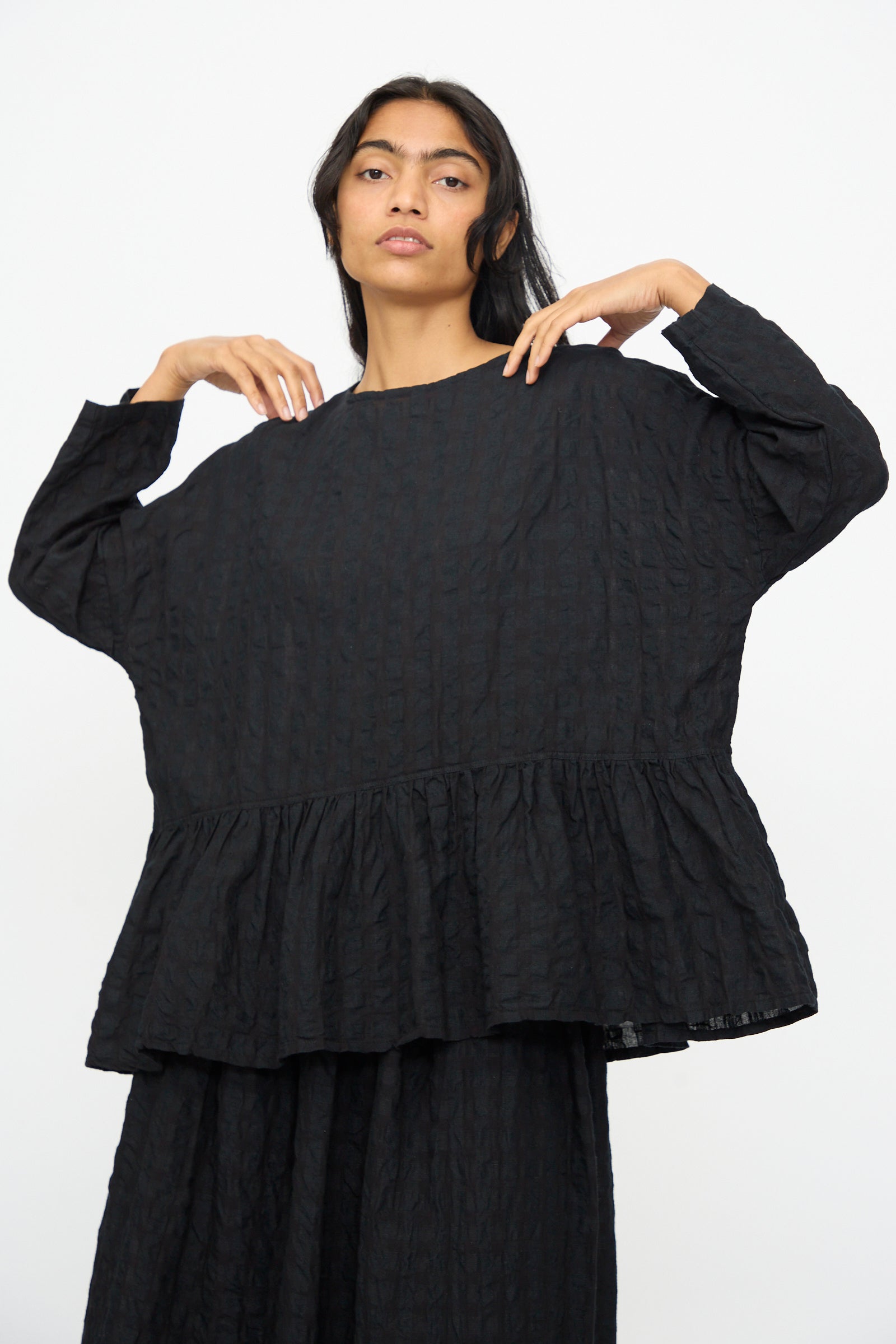 A person models the Ichi Antiquités Gingham Check Pullover in Black, a relaxed fit blouse with a cotton-linen blend and ruffled hem, standing against a plain white background.