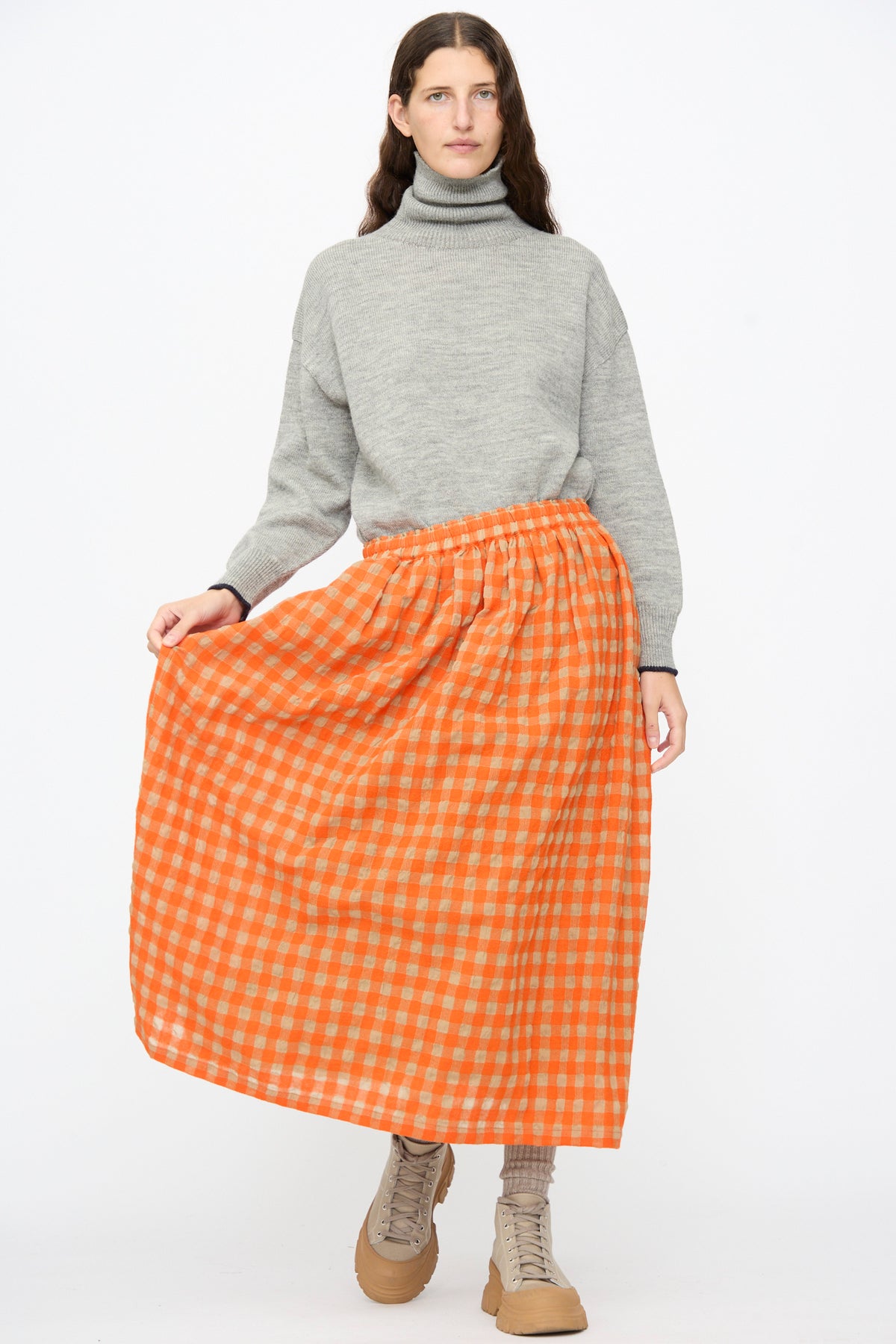 A model wearing a grey turtleneck sweater, an Ichi Antiquités Gingham Check Skirt in Beige and Orange, and beige lace-up boots.