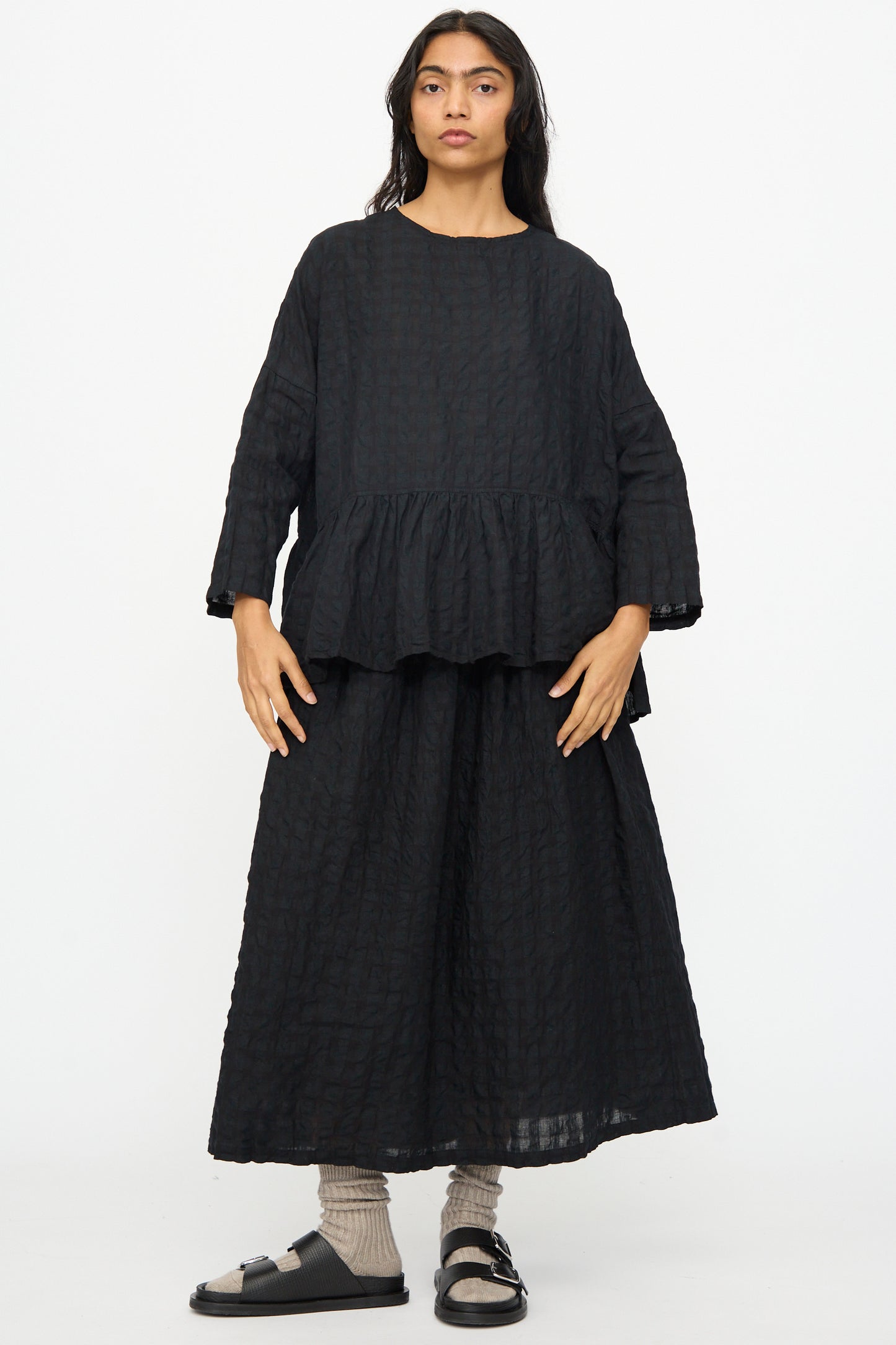 Person wears a loose-fitting black textured top with peplum hem, paired with the Ichi Antiquités Gingham Check Skirt in Black, gray socks, and black sandals, standing against a plain white background.
